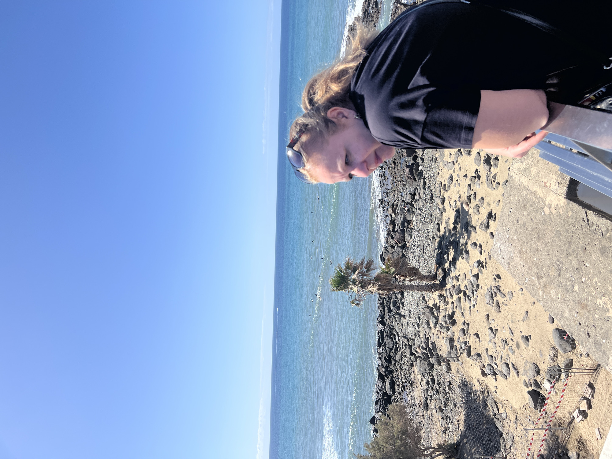 A medium shot captures a person leaning over a railing, gazing downward toward a beach. The beach consists of black volcanic rocks and coarse sand, fronting the ocean. Visible in the water are several birds. In the background, the cloudless blue sky meets the ocean's horizon. The scene appears to be in Maspalomas, Spain, judging from the volcanic rock. A barrier fence is visible near the beach's edge.