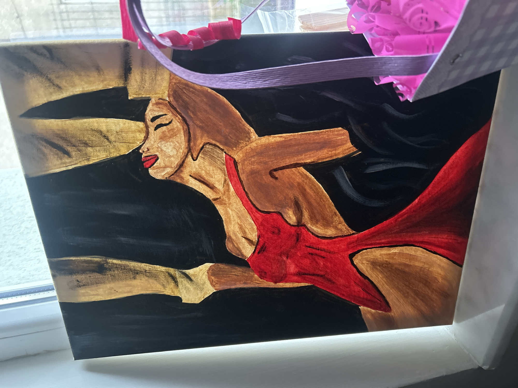 The image depicts a framed painting leaning against a window frame. The painting features a woman with tan skin, brown hair, and red lips, wearing a red garment. She is depicted against a background of black and gold horizontal strokes. Above the painting are various ribbons and decorative elements, including pink and purple materials. The painting and its frame are resting on a white window sill, with a window and blurry outdoor view visible in the background. The location in Plovdiv, Bulgaria, is not discernible from the image.