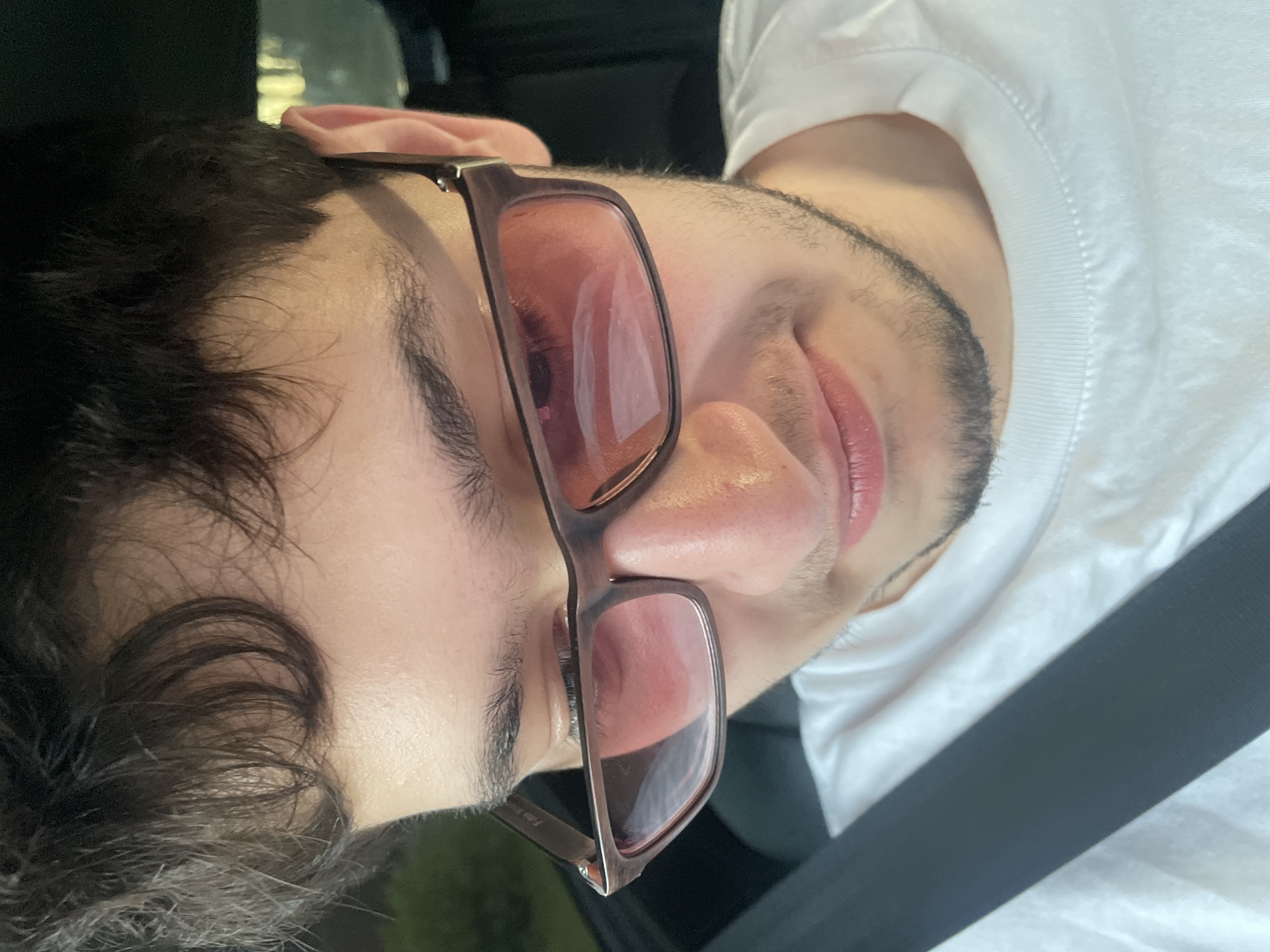 The image shows a close-up, head-and-shoulders view of a young man wearing a white t-shirt and dark-rimmed glasses with pink lenses. He has dark, curly hair and is looking downwards, with his face partially obscured by the glasses and his hair. A black seatbelt is visible across his chest. The background is dark and out of focus, with some blurry green foliage and a hint of light in the distance, suggesting an outdoor or car interior setting. There are no discernible landmarks or indicators to place this image specifically within Votkinsk, Russia.