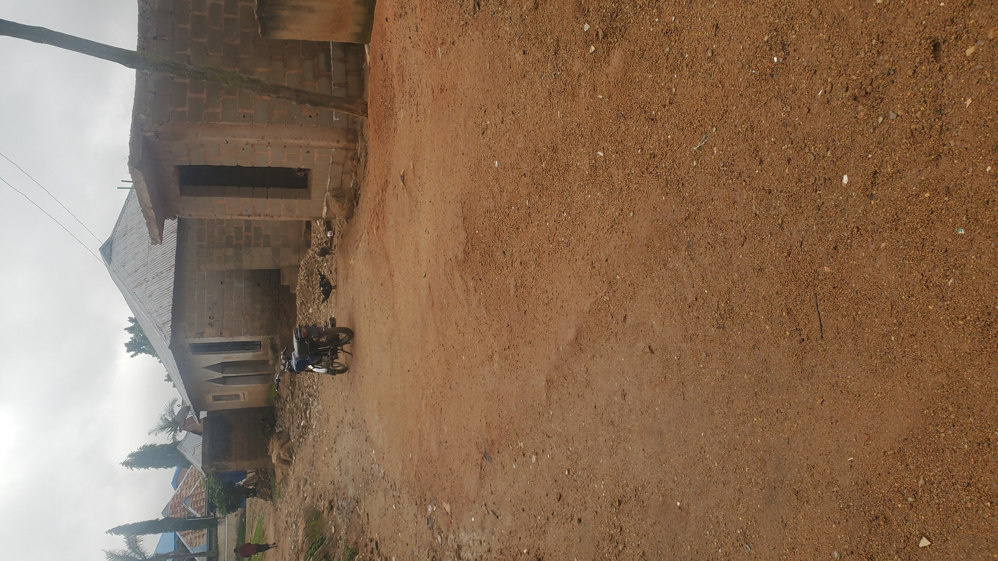 An outdoor, eye-level, medium shot captures a dirt road in front of two unfinished brick buildings under a cloudy sky. The building on the left has a corrugated metal roof and appears to be a home, with two arched doorways visible. The building on the right is in a more rudimentary state of construction, with an open doorway and visible concrete block walls. A motorcycle is parked on the road in front of the buildings, and several chickens are pecking at the ground nearby. A person in a red shirt is walking away from the camera on the left side of the road, and palm trees are visible in the background. This scene is consistent with a residential area in Jos, Nigeria, likely in a developing or rural part of the city where construction is ongoing.