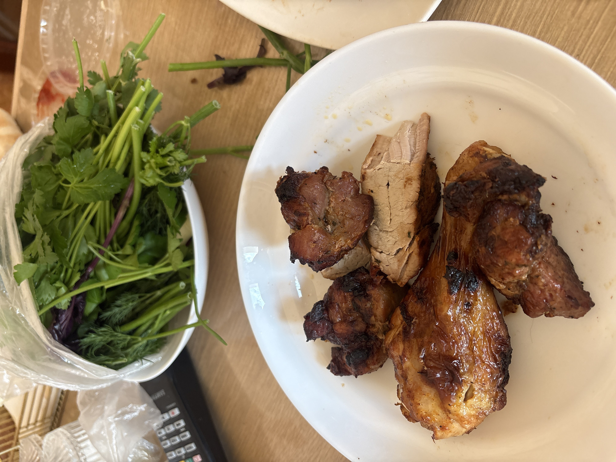 This image captures an overhead view of a meal served on a light wooden table, likely indoors, in Krasnoyarsk, Russia.
The main subjects are food items. On a round white ceramic plate, occupying the right side of the frame, several pieces of grilled meat are arranged. The meat exhibits a dark, caramelized exterior with some charred sections, indicative of high-heat cooking, potentially shashlik or similar grilled cuts. Some pieces are sliced, revealing lighter, cooked interior meat.
To the left, a white bowl holds a fresh assortment of herbs, partially encased in a clear plastic bag. Bright green cilantro and feathery dill are clearly visible, along with stems of purple basil, providing a vibrant green and purple contrast to the cooked meat.
Other elements in the scene include the textured wooden table surface, a crumpled clear plastic bag positioned at the bottom left, and a dark electronic device, possibly a remote control or calculator, with several unreadable buttons, peeking into the bottom left corner of the frame.
The image suggests a moment before or during a meal, with the food presented for consumption. No people are visible, and no specific activities are shown beyond the implied act of dining. The lighting is soft and even, typical of an indoor setting. No particular emotions are conveyed, and no legible text appears on any object within the frame.