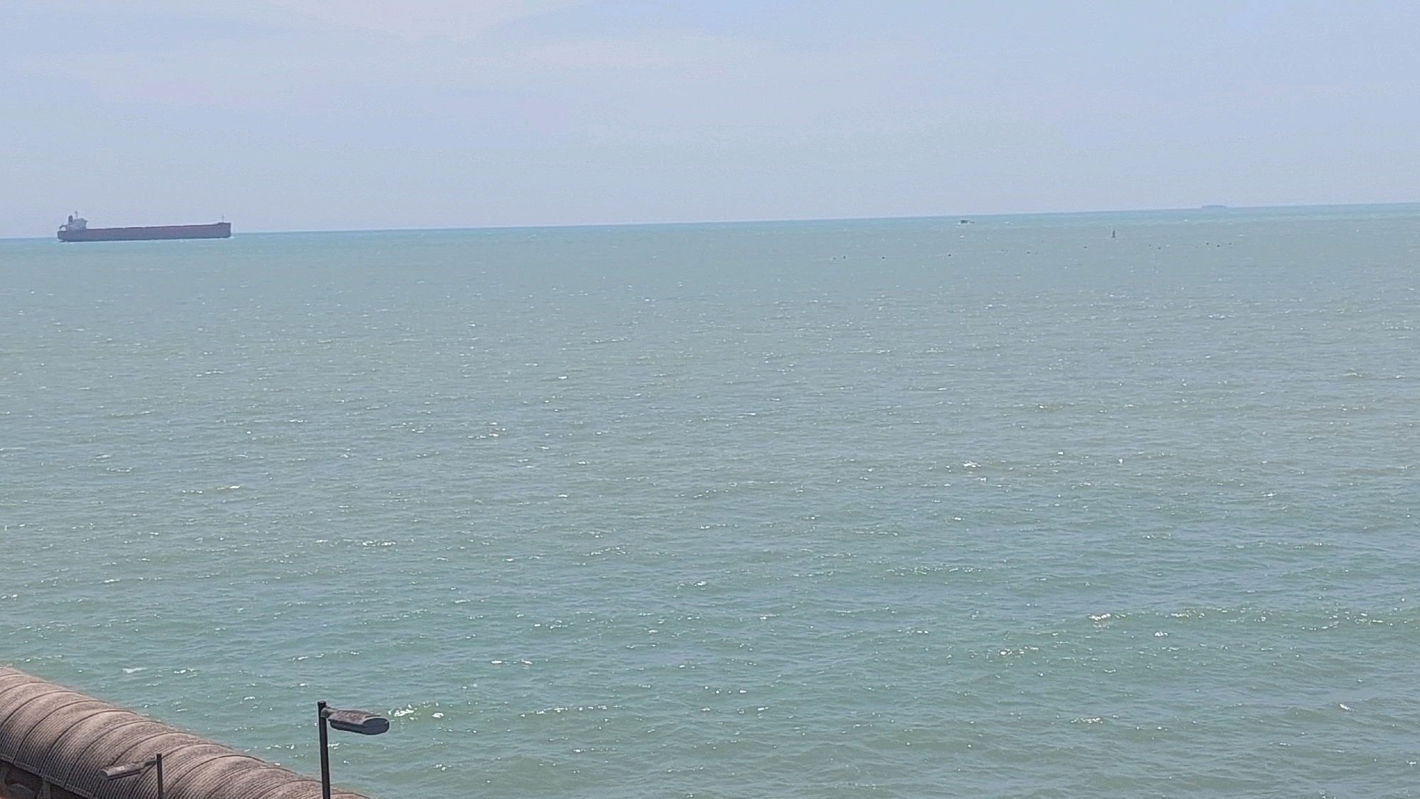 A wide-angle, eye-level shot captures a serene seascape under a pale sky. In the distance, a large, reddish-brown cargo ship is visible on the horizon, moving left to right across the frame. The sea is a muted turquoise, with gentle ripples and occasional glints of sunlight reflecting off the surface. The sky is a hazy, light blue, blending seamlessly with the horizon line. In the foreground, a textured, curved structure runs along the bottom of the image, appearing to be part of a seawall or embankment. A black lamppost with a modern, rectangular light fixture stands on this structure, pointing towards the sea. Another lamppost is partially visible to the left. The overall impression is one of tranquility and vastness, with the man-made elements contrasting with the natural expanse of the ocean.
