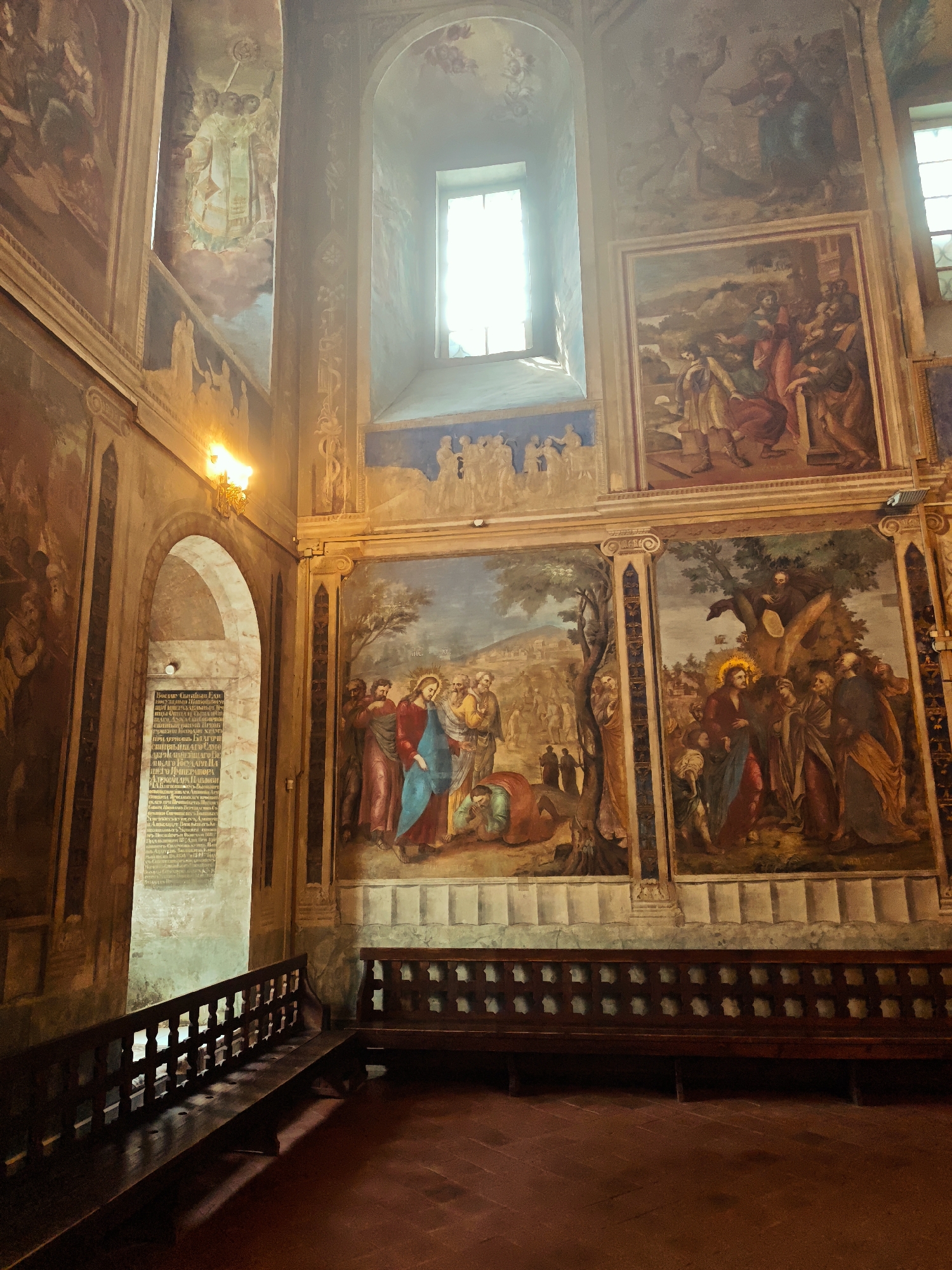 The image captures the interior of a historic building, possibly a church or chapel, characterized by religious paintings and a sense of reverence. The scene is dominated by large murals depicting biblical scenes or religious figures on the walls. A set of wooden benches are present, suggesting a space for prayer or reflection. A decorative wooden rail separates the seating area from an adjacent corridor where a plaque with Cyrillic text can be seen. The architectural details such as arched doorways and window frames, contribute to the building's historical character. The overall atmosphere is dim, with lighting provided by wall-mounted fixtures and natural light entering through a high window. While the location is noted as Fedotovo, Russia, the image itself is the main subject.