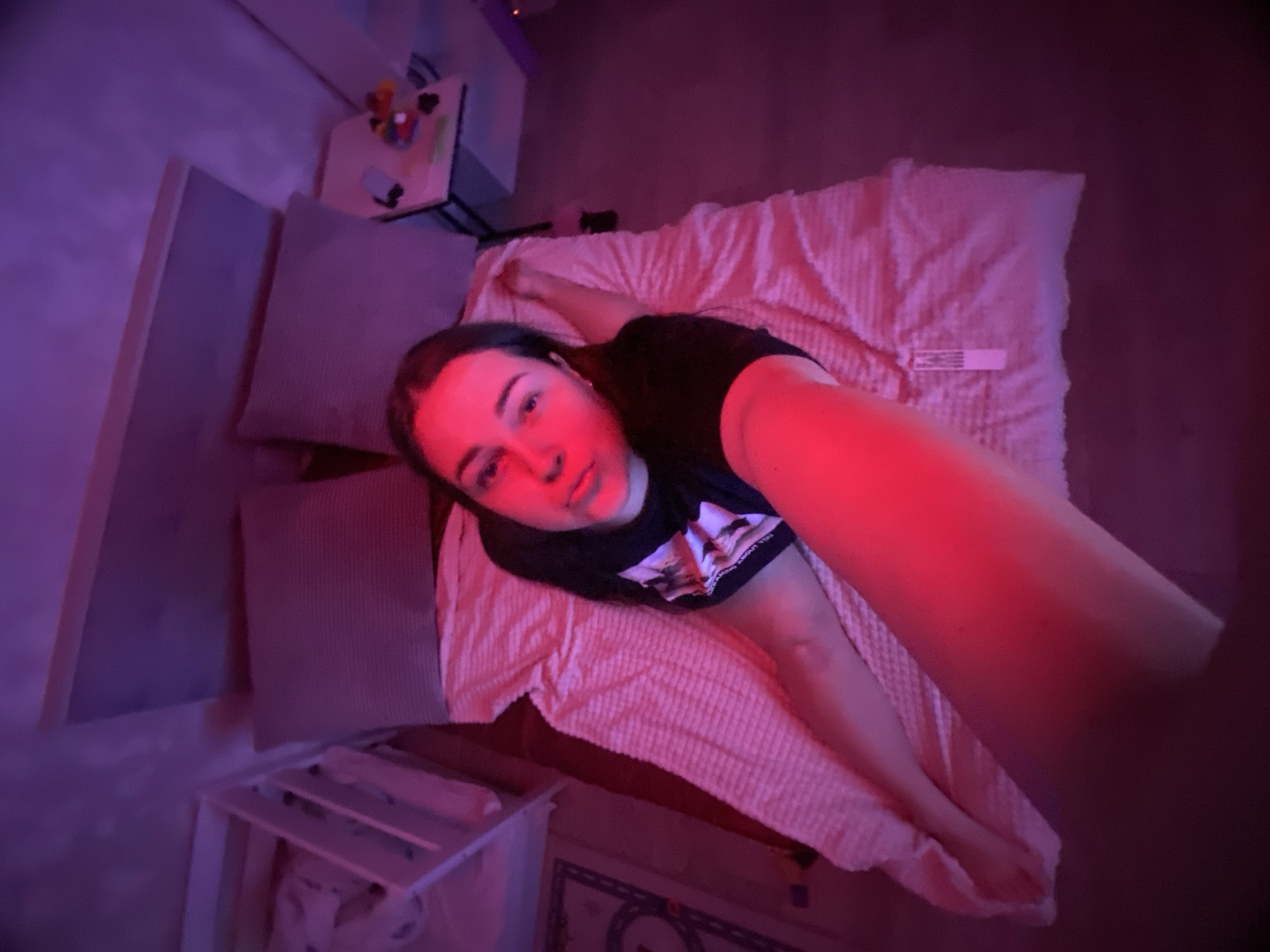 The image is a high-angle selfie featuring a young woman lying on a bed in an indoor setting, likely a bedroom in Sochi, Russia. The room is dimly lit, dominated by strong artificial red and purple lights, suggesting it is nighttime or a low-light environment.
The main subject is a young woman with dark hair, wearing a dark t-shirt with a graphic design and some indistinct white text below it. She is lying on her back on a bed, looking directly at the camera. Her right arm, which holds the camera to take the selfie, is extended into the foreground and is intensely illuminated by the red light, casting a vibrant red hue across her arm, face, and parts of the bed. Her expression appears neutral.
The bed is covered with a light-colored, textured blanket or duvet, which takes on a pinkish tint from the lighting. A small white rectangular tag with unreadable text is visible on the blanket. Behind her head, two grey pillows are propped against a headboard or wall, which is bathed in purple light.
To the left of the bed, a small side table or shelf is visible, holding various items including what appears to be a phone and some small, colorful objects. Below the bed, a white slatted shelf or rack can be seen, and on the floor, a mat or rug with a geometric pattern is partially visible. The overall mood of the scene is intimate and calm, created by the soft, colored lighting.