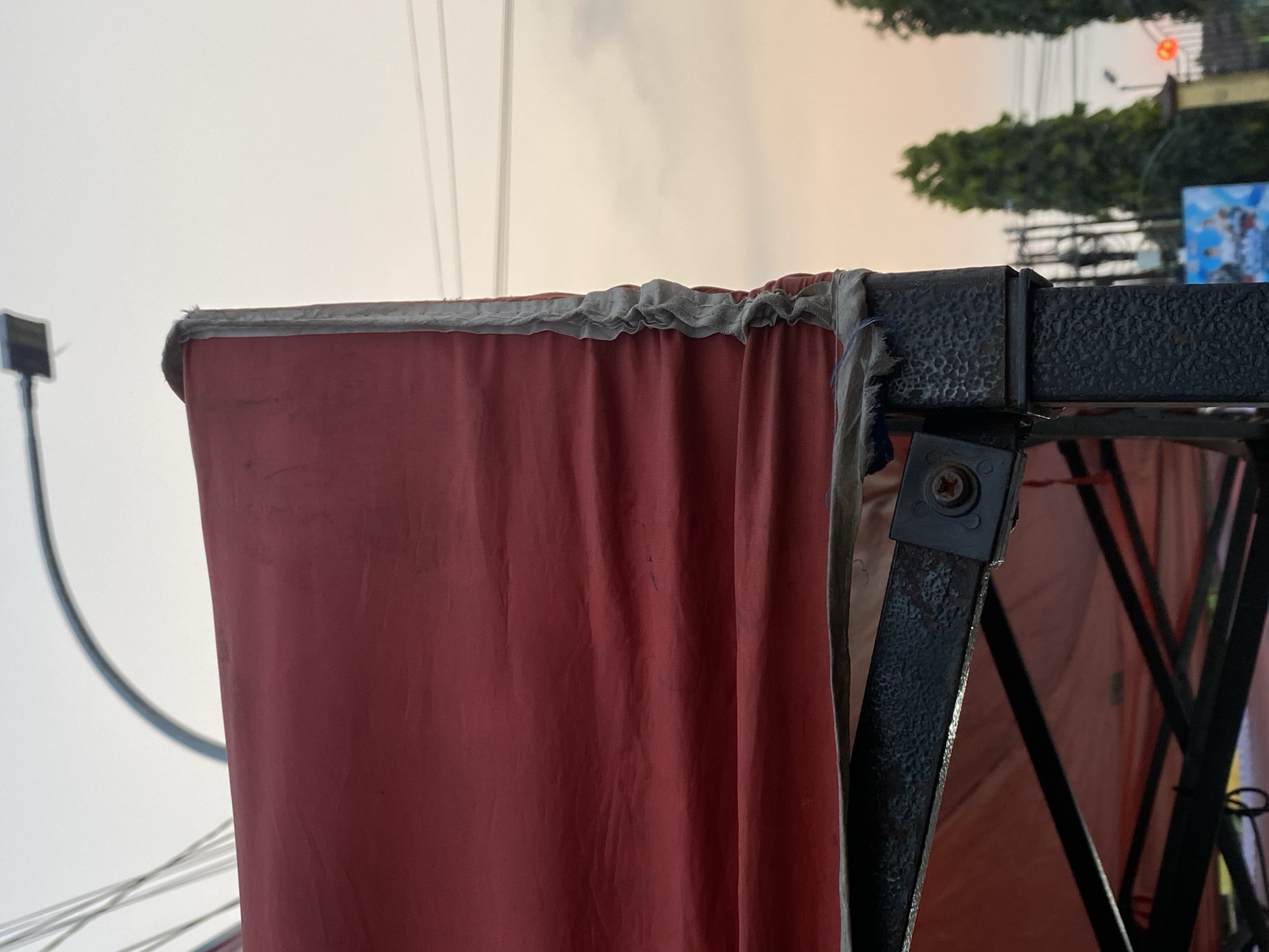 The image shows a close-up view of a red canopy of a portable stall, with a dark metal frame visible beneath. The canopy's edge appears worn and somewhat frayed, secured to the frame with a light-colored fabric trim. In the background, the sky is a soft orange and pink hue, suggesting either sunrise or sunset. Electrical wires and a street lamp are visible against the sky, along with silhouetted trees and what appears to be a building or fence in the distance. The overall impression is an outdoor urban scene, possibly in Kasihan, Indonesia, given the context.