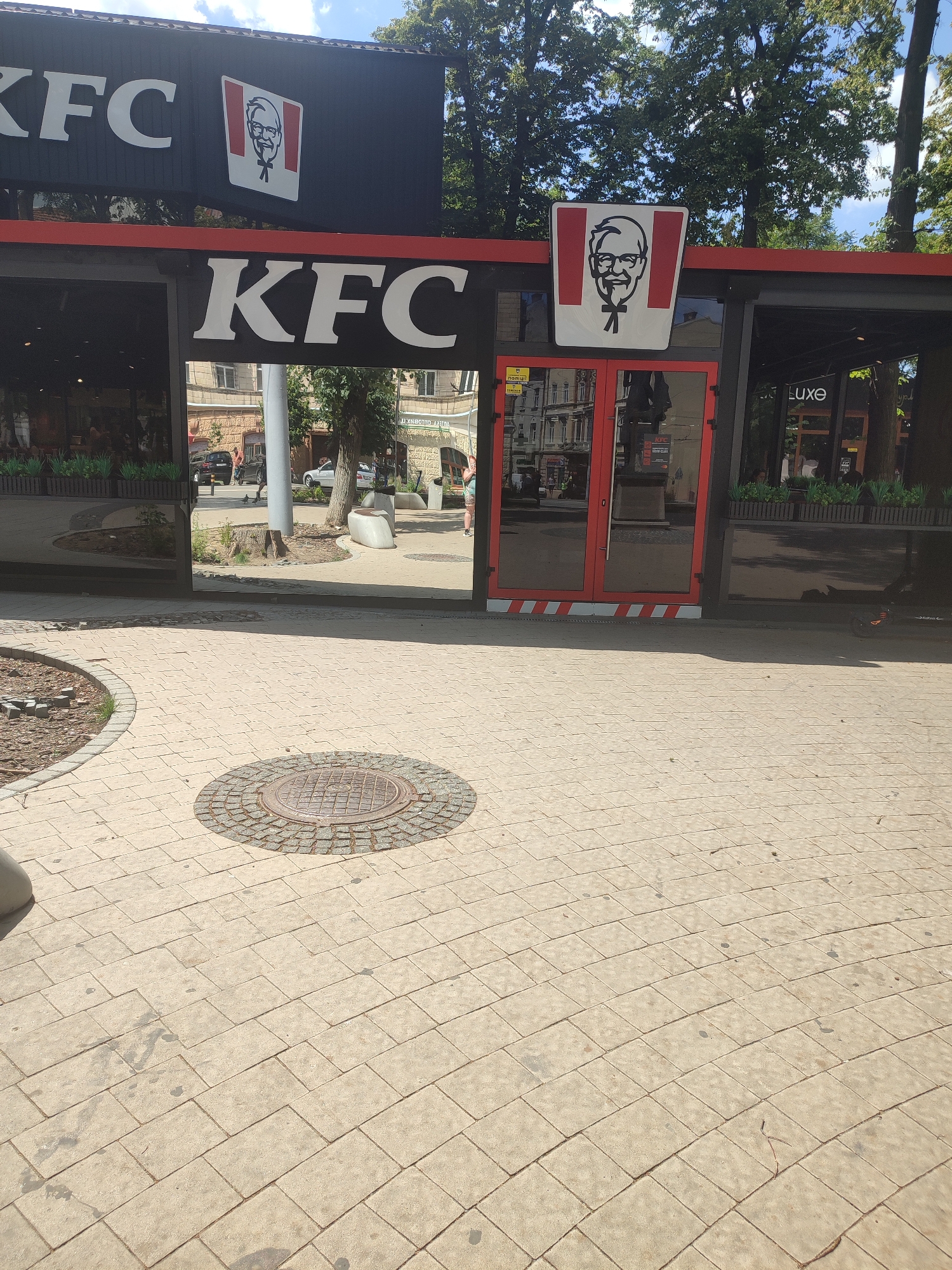 The image shows a KFC restaurant in Chernivtsi, Ukraine. The restaurant has a large KFC logo above the entrance, and another KFC logo on the side. There are also two red doors with glass panels. The pavement in front of the restaurant is made of beige tiles, and there is a manhole cover in the middle of the pavement. In the background, there are trees and buildings. The sky is blue and there are some clouds. There is a person walking in the distance, and a few cars parked on the street. The overall scene is a typical street view of a city in Ukraine.