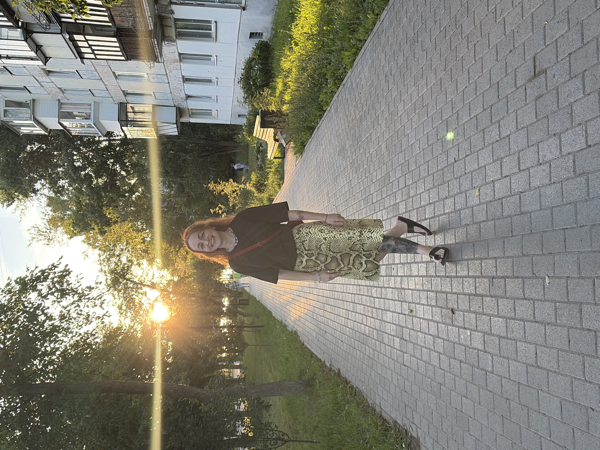 A woman with long, wavy red hair and a smiling expression stands on a paved walkway in Chaikovsky, Russia, during sunset. She is wearing a dark t-shirt, a light green snakeskin-patterned skirt, and dark sandals. Her left leg is visible, adorned with tattoos. A red strap from her bag is visible across her body. The sun is setting behind trees on the left side of the image, casting a warm, golden light and creating lens flare. On the right side of the walkway, a light-colored, multi-story apartment building is visible, with balconies and windows. Green grass and scattered bushes are present between the walkway and the building. The walkway is constructed of gray rectangular pavers. In the distance, other people are walking along the path.