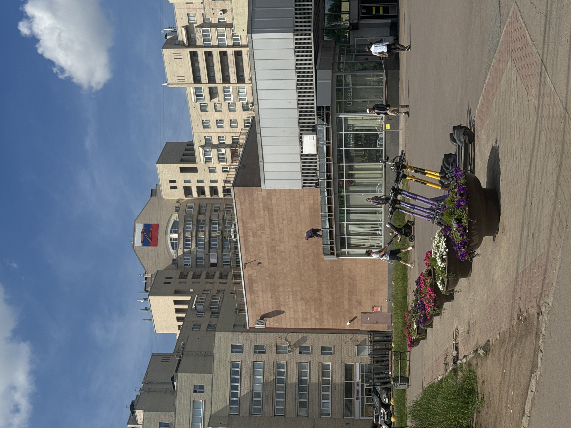 A view of a modern residential area in Saint Petersburg, Russia, on a sunny day with a blue sky and scattered clouds. The foreground features a paved walkway lined with large planters overflowing with colorful flowers, including pink, purple, and white petunias. Several electric scooters are parked in a row behind the planters.
Beyond the walkway, there is a light brown brick building with large windows and a glass-fronted section. A person in dark clothing is standing on a ledge near the top of this building. Further back, a complex of multi-story apartment buildings with beige facades dominates the background. A large Russian flag is prominently displayed on the facade of one of the taller buildings.
In the middle ground, several people are visible. One person is pushing a stroller, another is walking in the opposite direction, and two individuals are standing near a building entrance. Cars are parked in front of another building on the left side of the image. The environment appears clean and well-maintained.