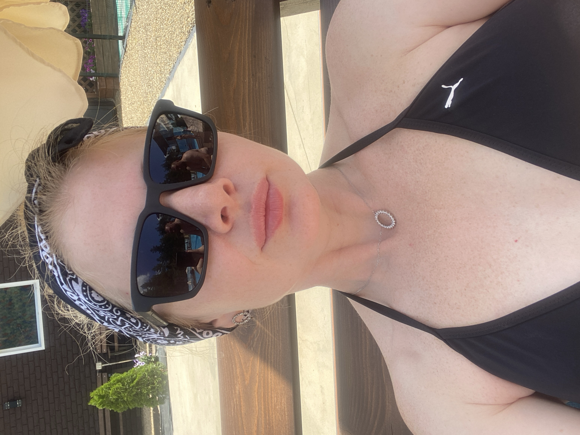 A woman with blonde hair tied back with a black and white paisley bandana is taking a selfie outdoors. She is wearing black sunglasses and a black bikini top with a Puma logo. A silver necklace with a circular pendant is visible. The background shows a building with a dark brick facade and a white-framed window, a green coniferous tree, and a body of water that appears to be a pool. The ground is covered with gravel. The scene appears to be a residential area, possibly a backyard or a resort. The weather is sunny.