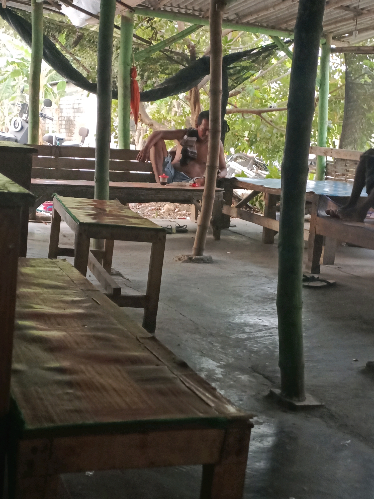 A person sits on a wooden bench in a covered outdoor area. The person is wearing a horizontally striped shirt and blue shorts. They are looking down. A small red cup is on the table in front of them. The area is supported by bamboo poles and has a corrugated metal roof. In the background, there are trees and a motorcycle. The ground is concrete. The scene is located in Rancang Kencono, Indonesia.