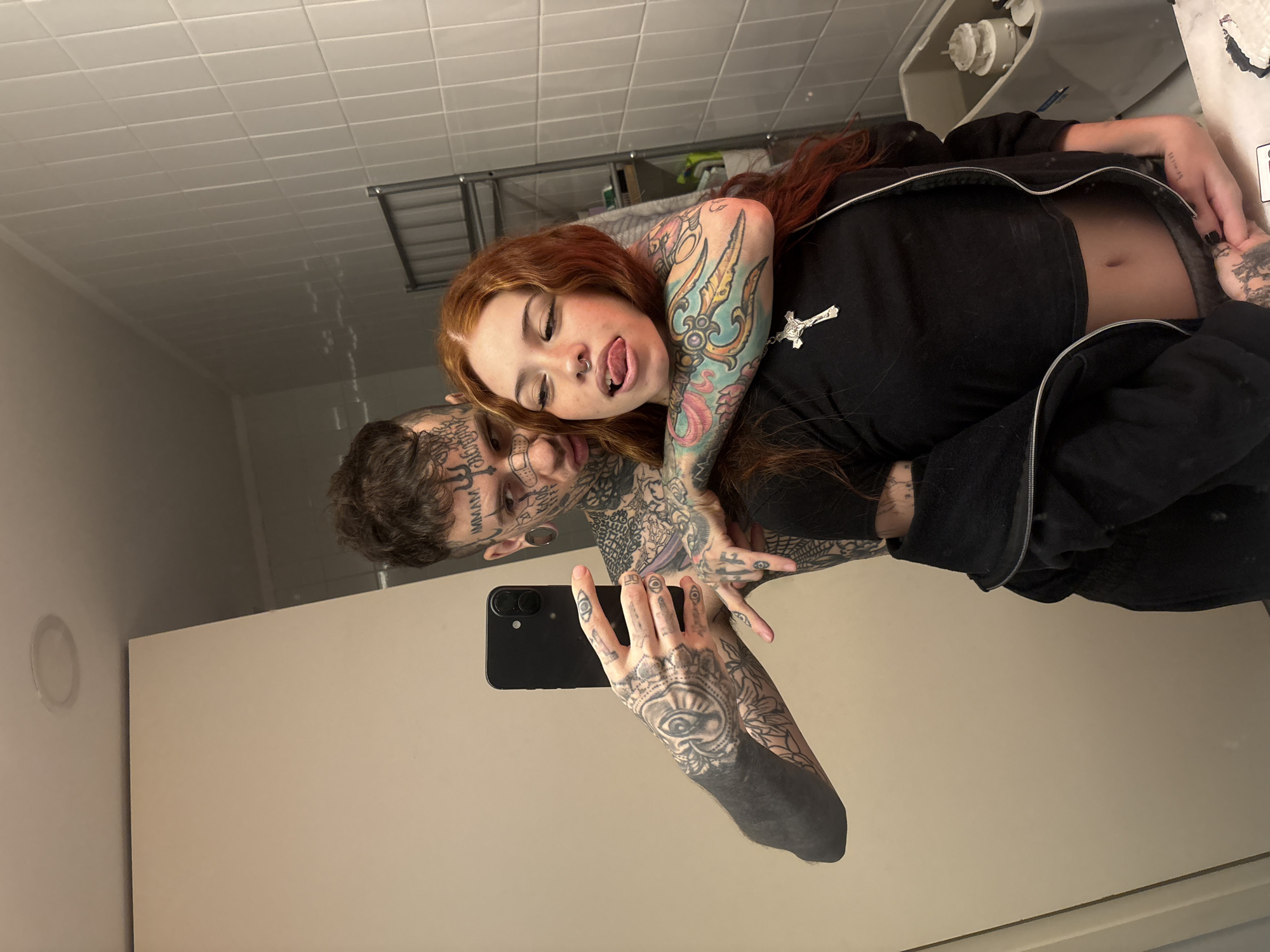 The image is a mirror selfie taken in an interior space, likely a bathroom, located in Buenos Aires, Argentina. Two individuals are depicted. A male subject, heavily tattooed across his face, head, neck, and arms (including "IMMAM" above his right eyebrow and "KAOS" below his left eye), with ear gauges, holds a black smartphone in his right hand to capture the image. He is shirtless. His left arm is positioned around the shoulders of a female subject, who has long reddish-brown hair, a nose piercing, and visible tattoos on her left arm. She wears a black cropped top, a silver crucifix necklace, and a black zip-up jacket draped over her shoulders. Her tongue is partially extended. The immediate background features white rectangular wall tiles, a metallic shelving unit, and the upper portion of a white toilet tank. A white ceiling with a circular recessed light fixture is visible at the top.