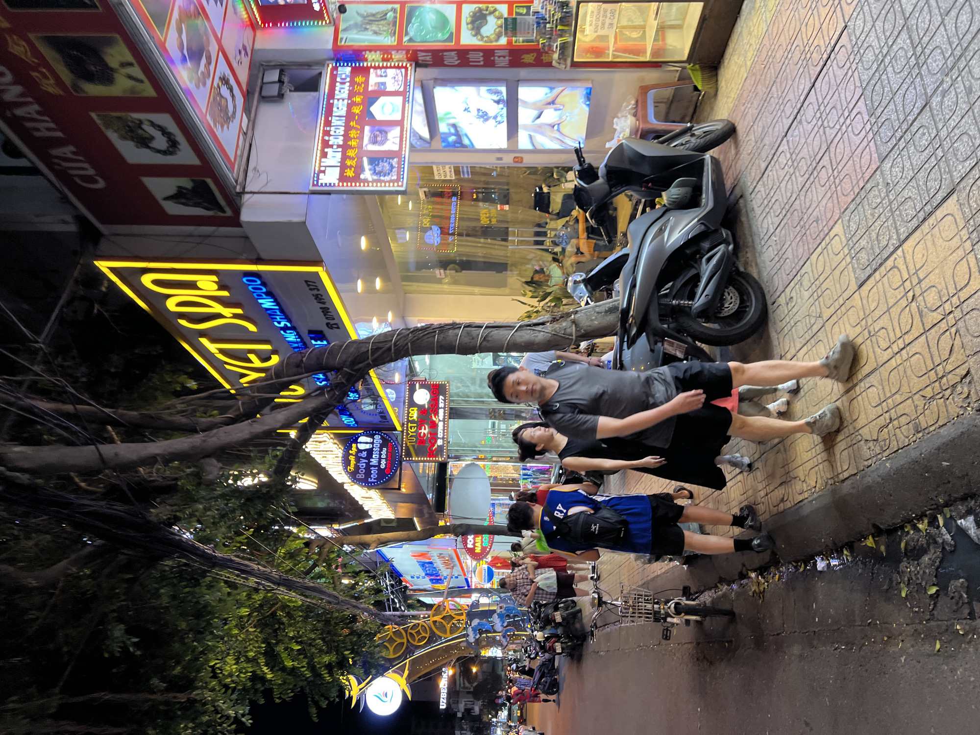 The image depicts a bustling street scene at night in Nha Trang, Vietnam. A sidewalk is lined with shops and businesses, illuminated by neon signs. Several people are walking along the sidewalk, some appearing to be tourists. Motorcycles and a bicycle are parked along the street. Trees with exposed roots line the sidewalk, casting shadows. One shop appears to be a spa or massage parlor, with a sign reading "Body & Foot Massage." Another sign indicates a "Mini Mart." The overall atmosphere is lively and active, typical of a commercial street in a Vietnamese city at night.
