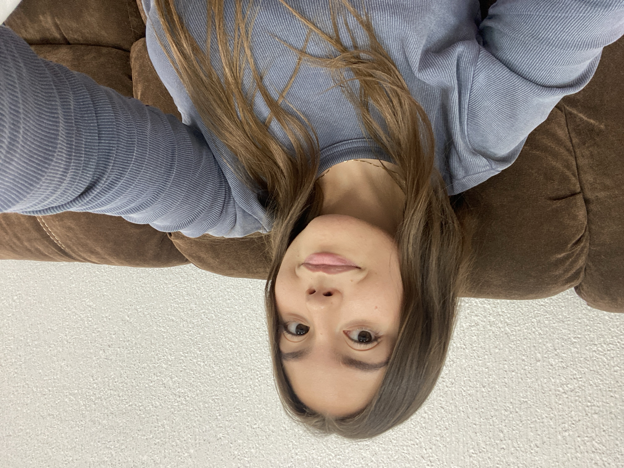 A young woman with brown hair is lying on a brown couch, wearing a blue long-sleeved shirt. The background appears to be a white textured wall. The location is unknown.
