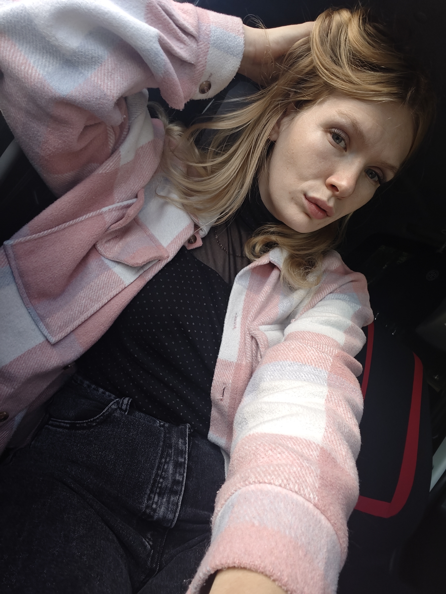 A young woman with blonde hair and blue eyes is sitting in a car. She is wearing a pink and white plaid jacket over a black sheer top and dark jeans. Her left hand is in her hair, and she is looking towards the camera. The car's interior is visible in the background, including a black seat with red trim. The lighting suggests it is daytime. The location is not specified in the image.