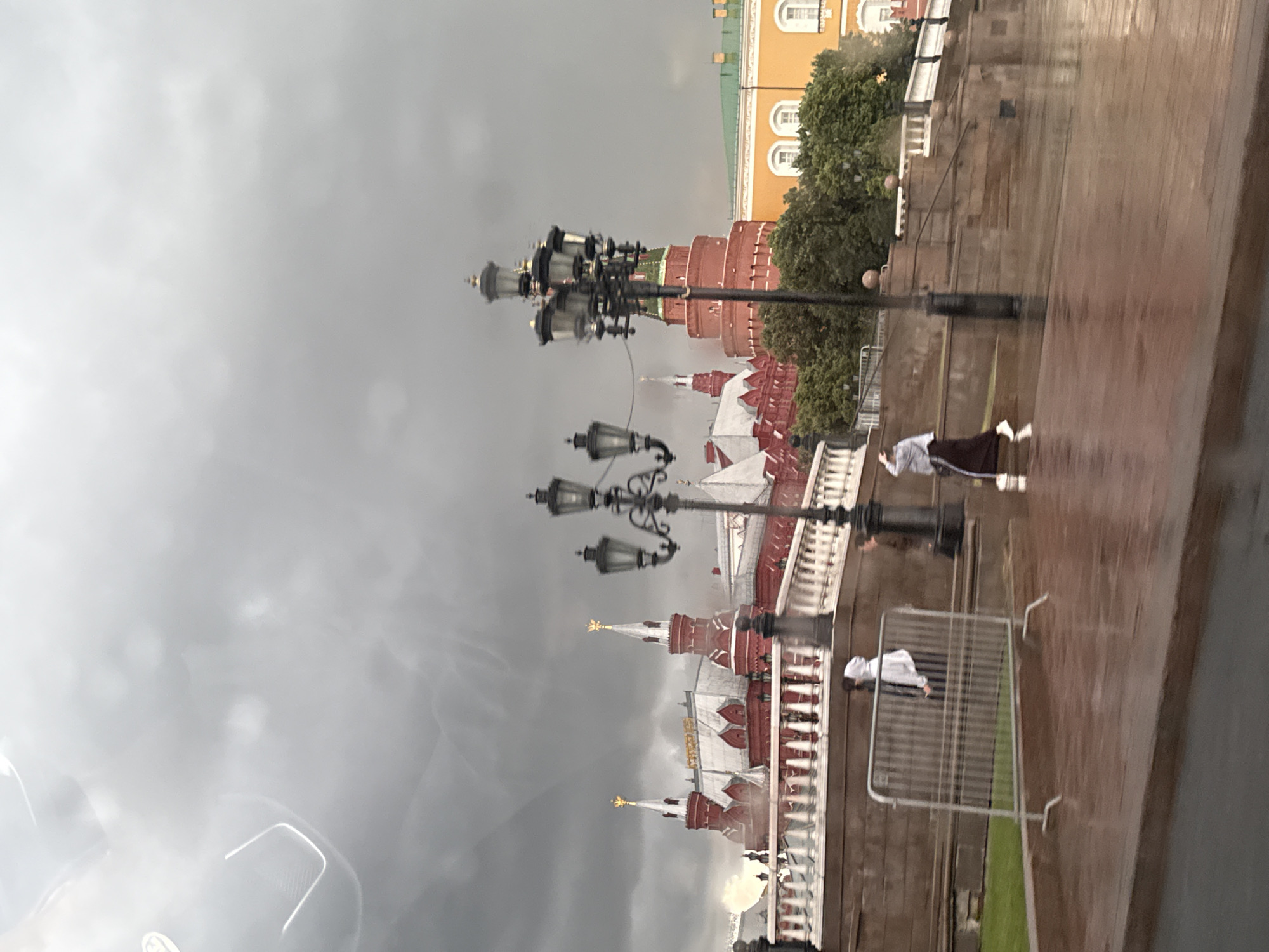 The media file captures a wet scene in Moscow, Russia, likely on a cloudy or rainy day. The prominent subjects include a series of ornate lampposts reflecting on the wet ground, and the distinctive red brick architecture of the Kremlin with its white and gold spires. The wet cobblestone ground creates strong reflections, blurring the details but suggesting a recent rainfall.
Two individuals are visible. One person, wearing a white hoodie, is standing near a metal barrier, appearing to be looking at the Kremlin. Another person, dressed in dark clothing with a white element on their head, is partially visible walking on the wet pathway.
The activity suggests a public space, possibly Red Square or an area adjacent to the Kremlin, experiencing a period of inclement weather. The atmosphere is somewhat somber due to the overcast sky and wet conditions, but the grandeur of the Kremlin architecture remains striking. There is no visible text in the media file. The time of day is not definitively clear but the lighting suggests it could be daytime, possibly late afternoon or early evening.