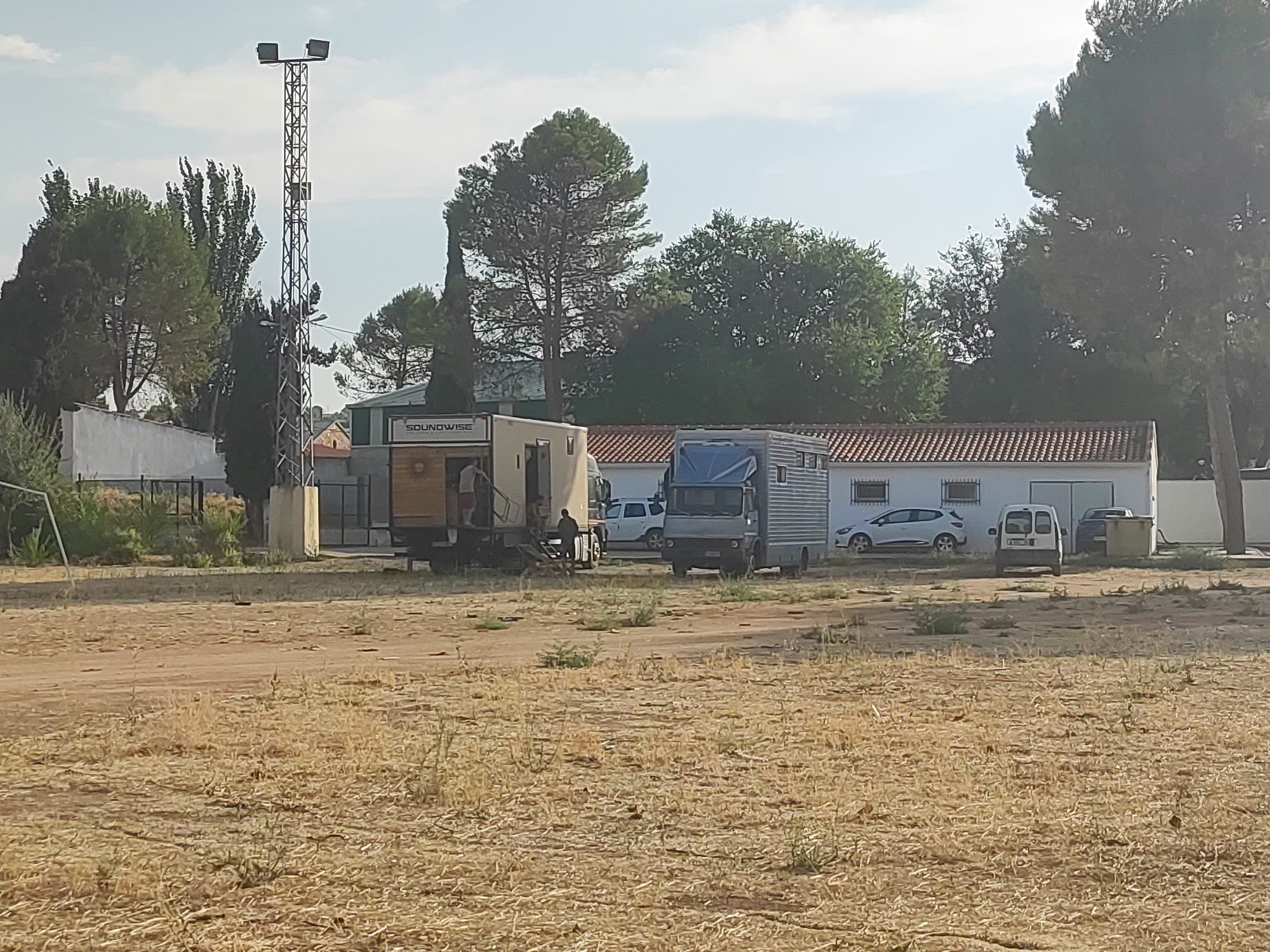 A group of vehicles, including a large truck and two trailers, are parked in an open, grassy field. The field appears dry and sparsely vegetated, with some areas of loose dirt. In the background, a low-rise building with a tiled roof is visible, and several trees are scattered throughout the scene. A tall, metal tower with lights stands to the left of the frame. There are a few people visible near the vehicles, some appearing to be interacting with the trailers. The sky is clear and blue, suggesting it is daytime. The overall impression is one of an outdoor event or gathering, possibly related to a traveling performance or a temporary setup.