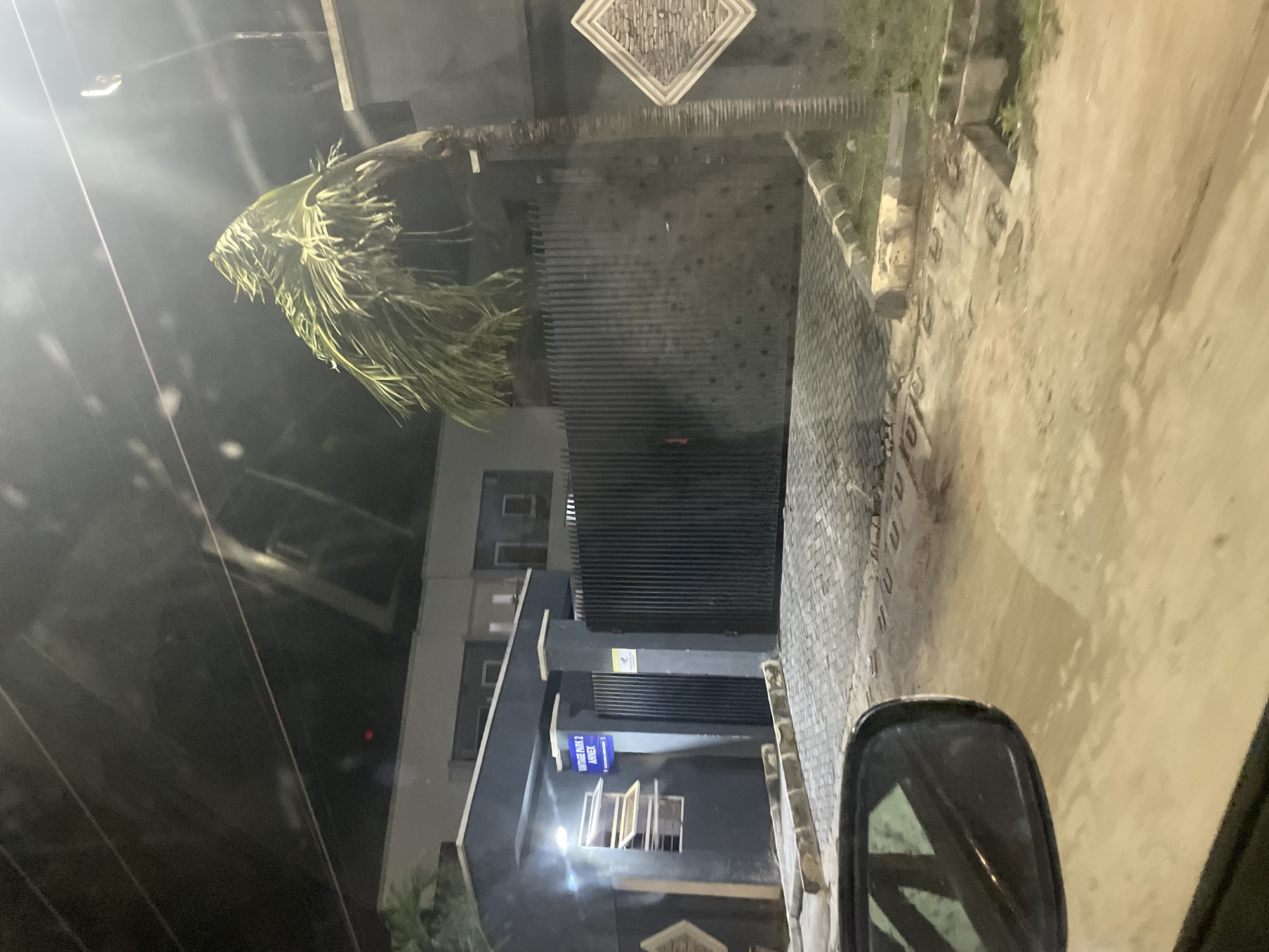 The image depicts a building in Igboefon, Nigeria, at night. A palm tree hangs over a gray brick driveway and sidewalk. The building has a dark gray facade with a sign that reads "VINTAGE BANK 2 ANNEX" on a blue background. The building has closed windows and a large black gate in front. A vehicle side mirror is visible in the lower right corner. The street appears wet.