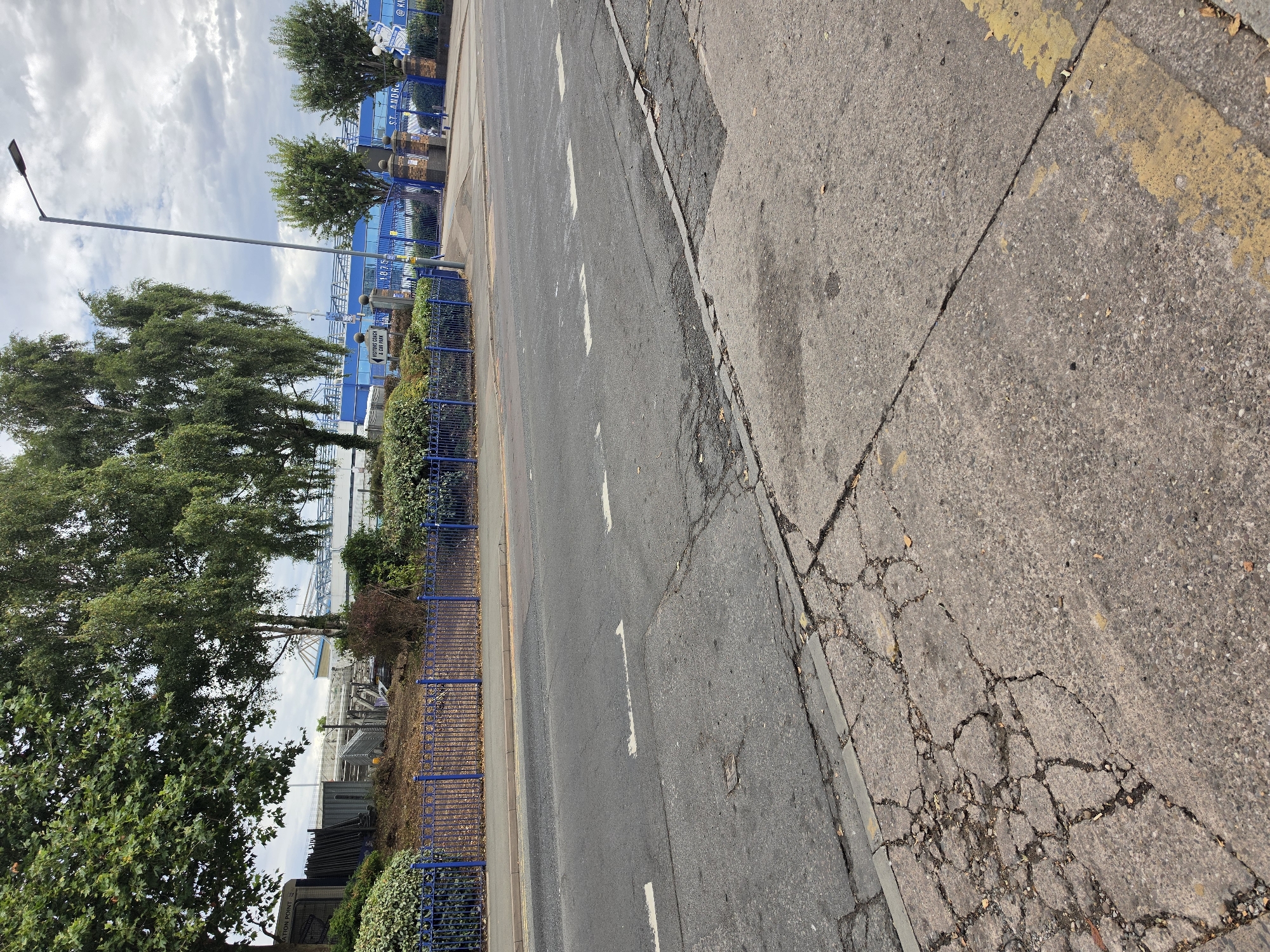 The image shows a view of the St Andrew's Stadium, home of Birmingham City Football Club, from across a road. The stadium is visible in the background with its blue seating and white roof structure. A blue metal fence runs along the front of the stadium grounds, separating it from the road. There are trees and bushes in the foreground and surrounding the stadium. A tall street light is visible on the right side of the image. The road in the foreground shows signs of wear and tear, with cracks and patches in the asphalt. Yellow lines mark the edge of the road. The sky is overcast with clouds. There are no people clearly visible in the scene, though some indistinct figures can be seen near the stadium entrance. The overall impression is of an urban environment on a cloudy day, with a focus on the football stadium.