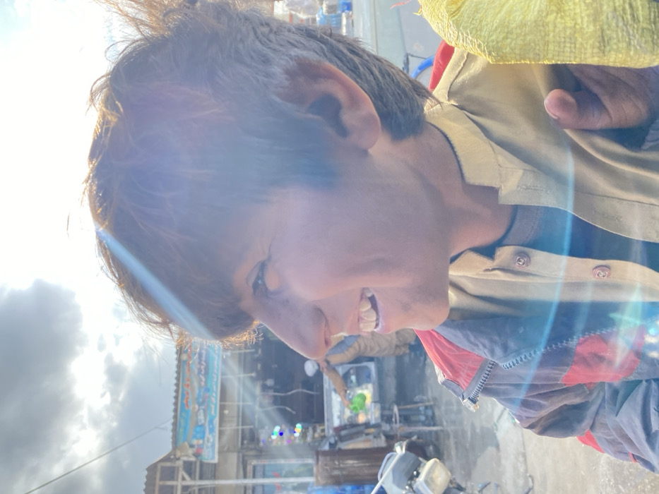 A young male is depicted in a right-profile view on a street in Kandahar, Afghanistan. He has short, dark hair, is smiling broadly with visible teeth, and is looking upwards and to the right. He wears a dark blue and red zippered jacket over a light tan, collared garment. His right hand holds a textured yellow object, resembling a bag. Bright sunlight from the upper left creates lens flare across the image.
In the mid-ground, to the left, a portion of a dark-colored motorbike or scooter is visible. Behind the subject, a commercial establishment with a corrugated metal roof and a light blue sign displaying white text (likely local script) is present. Below the sign, colorful lights (red, blue, green) are visible. Another individual, dressed in a light brown garment, is partially visible behind a counter within this establishment, holding a green object, possibly a pitcher or watering can. Various stacked transparent containers are visible in the background. The sky is partially cloudy.