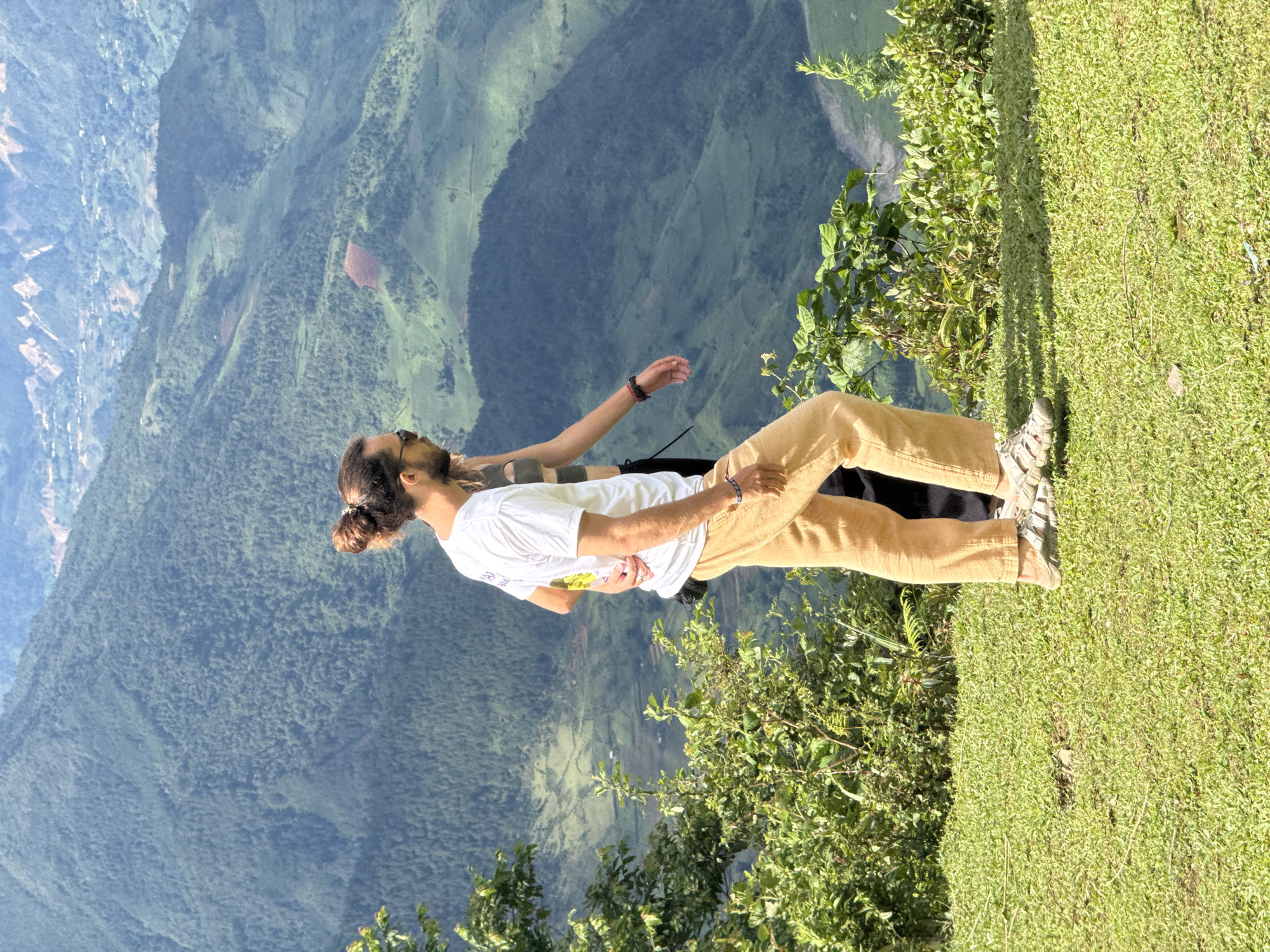The image captures two individuals on a grassy overlook in Tu Le, Vietnam, against a panoramic backdrop of green, terraced mountains. The primary subject, a male on the left, is depicted from mid-thigh up, facing right. He has dark hair tied in a bun, a beard, and wears sunglasses, a white t-shirt, light brown pants, and light-colored sandals. A blue bracelet is visible on his left wrist. His left hand rests on the lower back of a female partially obscured by his body, positioned to his right. The female is visible by her left arm and a portion of her torso, wearing what appears to be a dark, possibly striped, top. Both individuals are in motion, walking from left to right across the foreground's bright green, short-cut grass. Sparse green bushes are present on the left and along the edge of the overlook. The background is dominated by numerous layers of densely forested mountains and valleys, displaying varied shades of green, with some lighter patches indicating cultivation and reddish-brown sections of exposed earth. Faint, jagged mountain peaks are discernible in the far distance under a clear, bright sky. A river is subtly visible in the valley below, winding towards the right side of the frame.