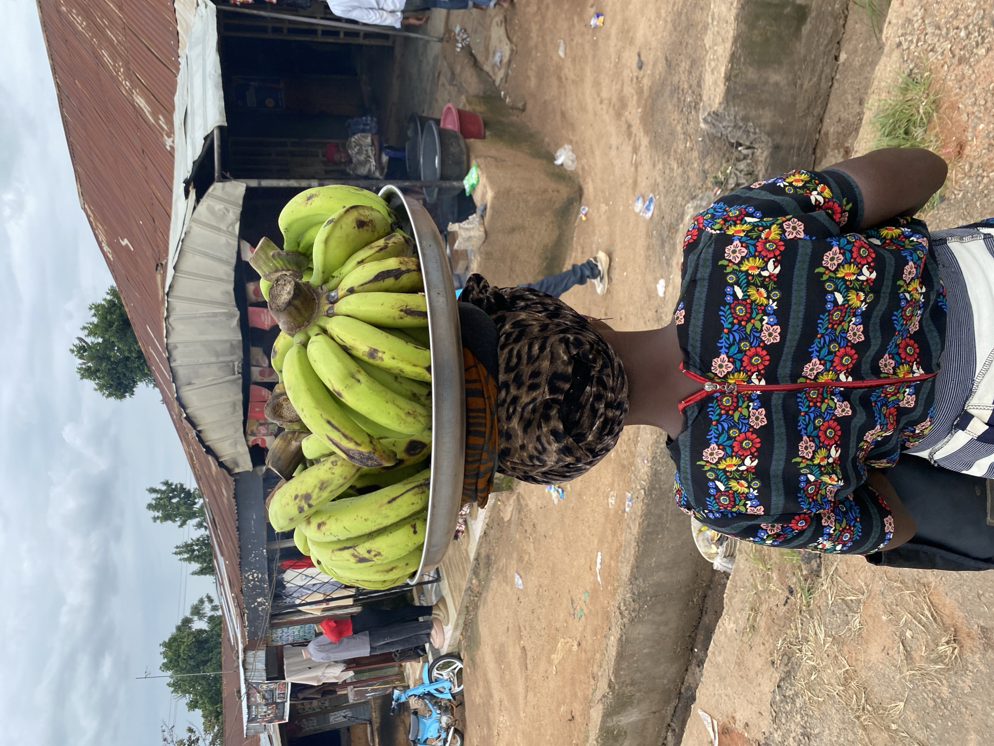 In Bukuru, Nigeria, a woman carries a large tray of green bananas balanced on her head. She wears a black, short-sleeved shirt with a colorful floral pattern and a red zipper, and a striped skirt. A leopard-print headscarf is visible beneath the tray. The background shows market stalls with corrugated metal roofs and various goods displayed, including clothing and what appear to be plastic containers. A blue motorcycle is parked to the left, and a few people are visible in the distance, some browsing stalls and others walking. The ground is dirt and gravel, with concrete drainage channels present. The sky is overcast.