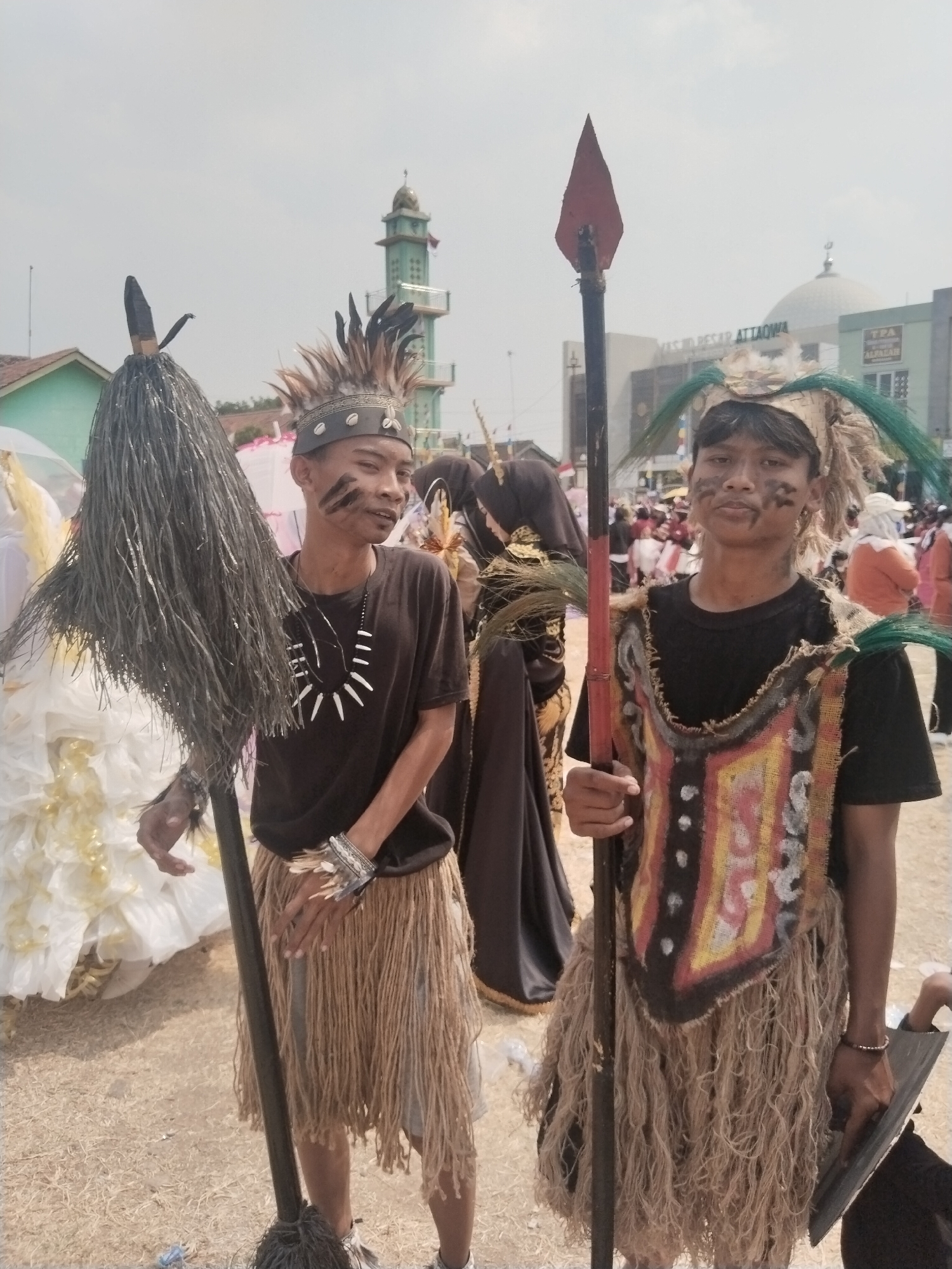 Two young men in traditional attire stand facing the camera in Margasari, Indonesia. The man on the left wears a black t-shirt, a necklace made of white shells, and a skirt of woven brown fibers. He holds a long staff topped with a dark, bristly object. His face is painted with black stripes. The man on the right wears a black t-shirt, a vest made of rough, colorful fabric, and a skirt of similar brown fibers. He also holds a spear with a red tip. He has black face paint as well. Behind them, a crowd of people and various structures are visible, including a mosque with a green dome and minaret. The ground is covered in dry grass and debris. The sky is overcast.