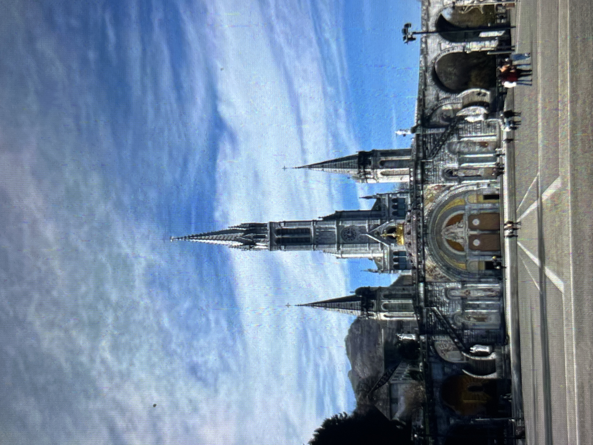 The image shows the Basilica of Our Lady of the Rosary of Lourdes in Tinaquillo, Venezuela. The basilica is a tall, ornate structure with multiple spires and arches. A mountainous landscape is visible in the background. In the foreground, there is a large paved area with white lines, and a few people are visible walking near the basilica. The sky is blue with streaky white clouds.
