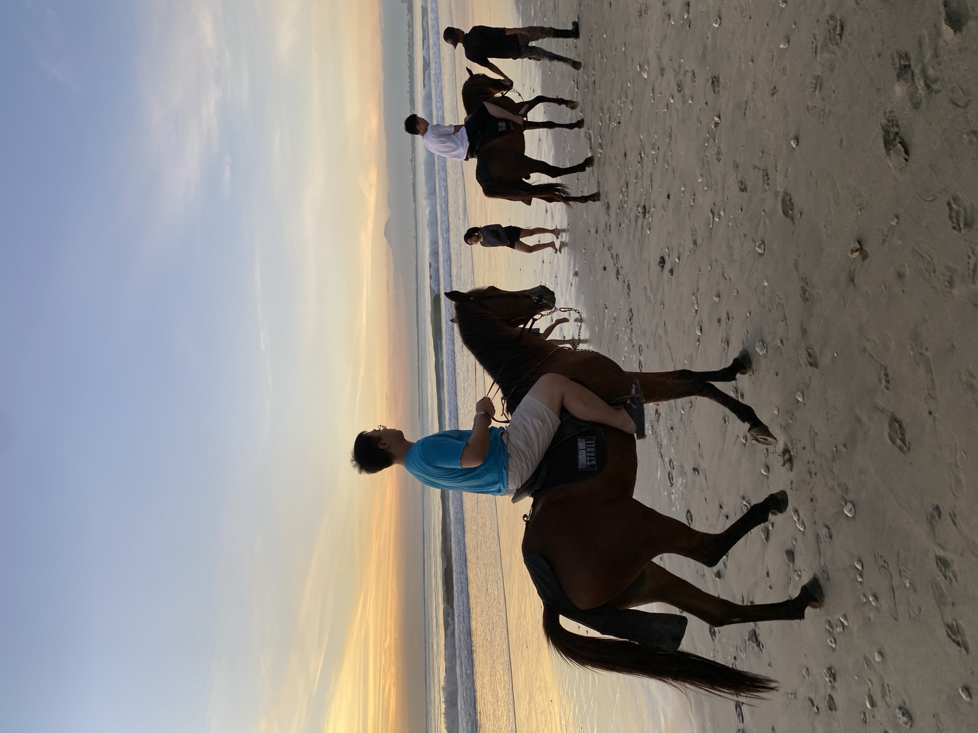 The image depicts four individuals riding horses on a beach at dusk in Kuta Selatan, Indonesia. The sky displays hues of blue and orange, indicating either sunrise or sunset. The sand is wet and reflective. The riders are positioned in a line, with one rider closer to the camera and the other three receding into the distance. All the horses are brown. The first rider is wearing a blue shirt and light-colored shorts. The second rider has a white shirt and dark pants. The other two riders in the distance are wearing dark clothing. The ocean is to the left of the riders.