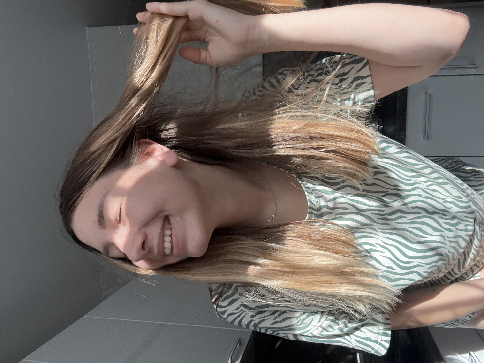 A young woman with long, blonde hair smiles broadly with her eyes closed as sunlight streams across her face. She is wearing a green and white patterned shirt. Her left hand is raised, touching her hair. The setting appears to be indoors, with a grey wall and tiled surface visible. The lighting suggests it is daytime, possibly morning or afternoon, with the sun creating strong highlights and shadows.