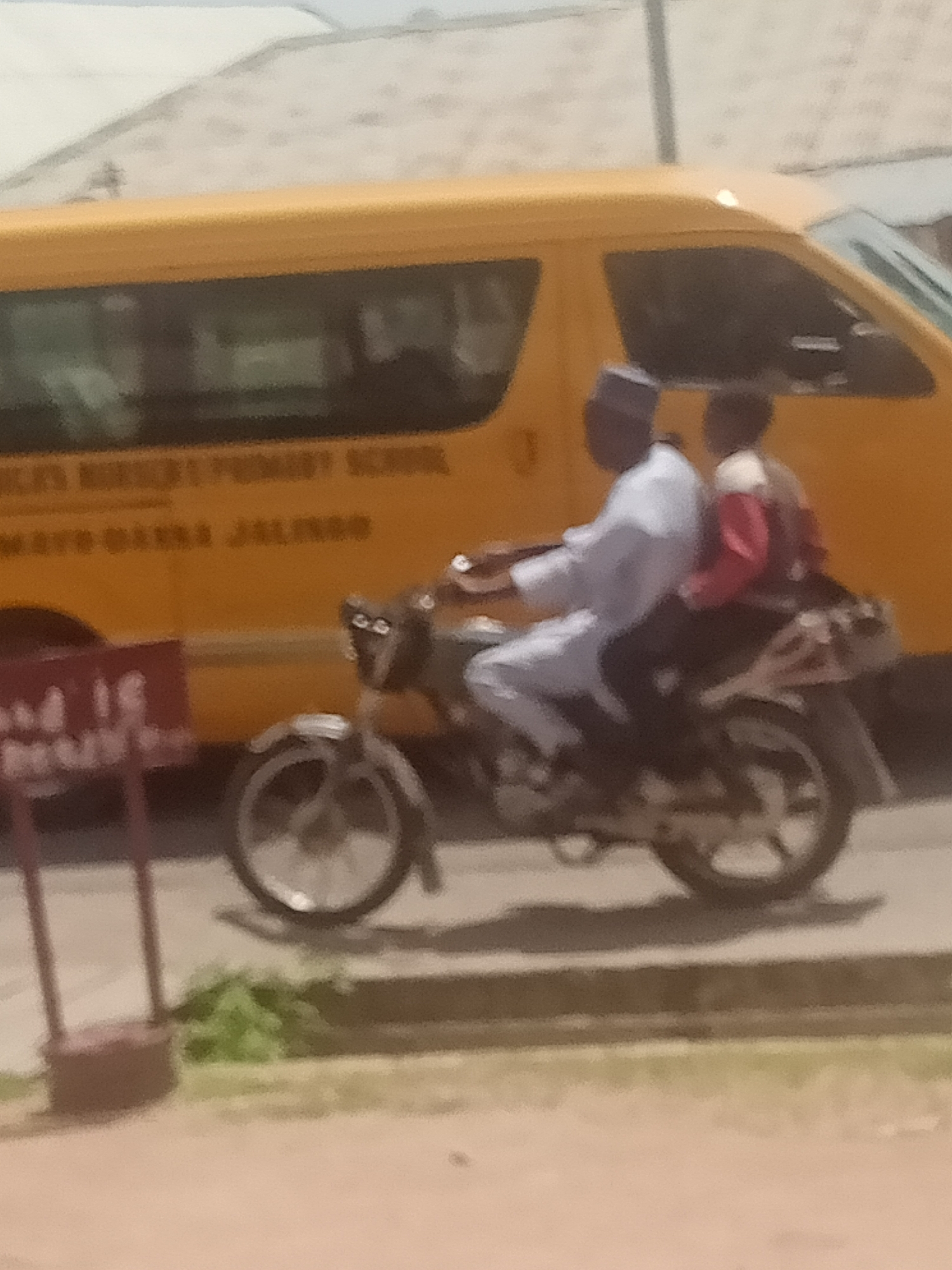 In Jalingo, Nigeria, two individuals are seen riding on a motorcycle. The person in the front is wearing a white robe and a cap, and is operating the motorcycle. The person in the back is wearing a red and white jacket. They are traveling alongside a yellow school bus with text on its side, which appears to read "MAYO RANSOM PRIMARY SCHOOL MAYO-BANNA JALINGO". A red sign on a post is visible to the left of the motorcycle. The scene is outdoors with a paved surface and some vegetation.