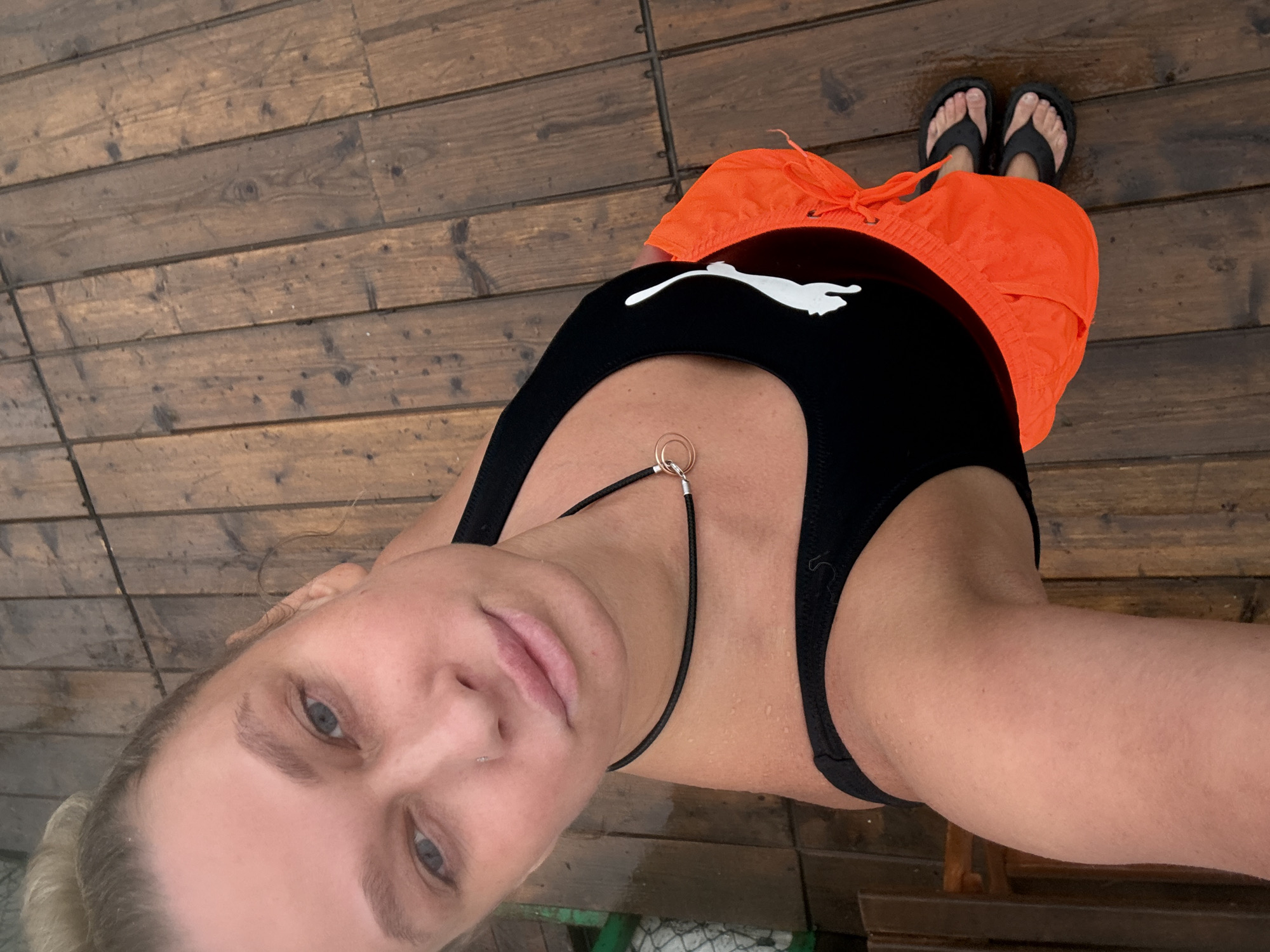 A fair-skinned female is seen from a high-angle, selfie perspective. She is wearing a black Puma branded sports bra, bright orange shorts, and black flip-flops. She also wears a thin black cord necklace with a gold ring. The backdrop is a wooden deck, possibly wet. The location is potentially Perm, Russia.