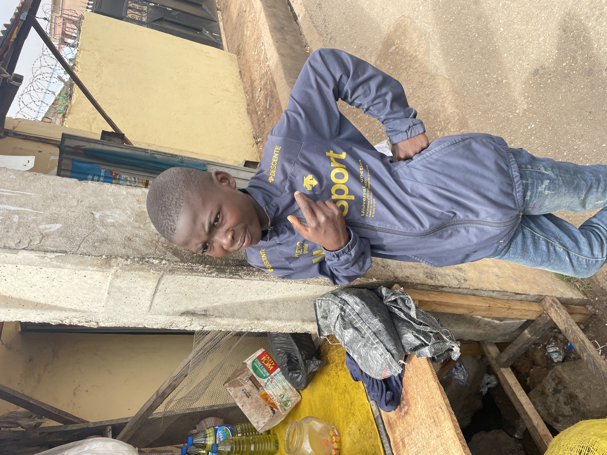 The image depicts a young man standing outdoors, possibly in a market or urban area in Jos, Nigeria. He is wearing a blue jacket with yellow lettering that includes the brand "Descente" and the word "sport." He's also wearing jeans. The man is making a hand gesture towards the camera. Behind him is a concrete wall with a yellow building visible in the background, topped with barbed wire. In the foreground, there is a makeshift table with various items, including cooking oil bottles, a package of "Tasty Tom" tomato paste, and what appears to be a jar of spices or other food items. Below the table are wooden supports revealing a dark space beneath. The scene suggests a potentially informal economic setting. The ground is a mix of concrete and dirt.
