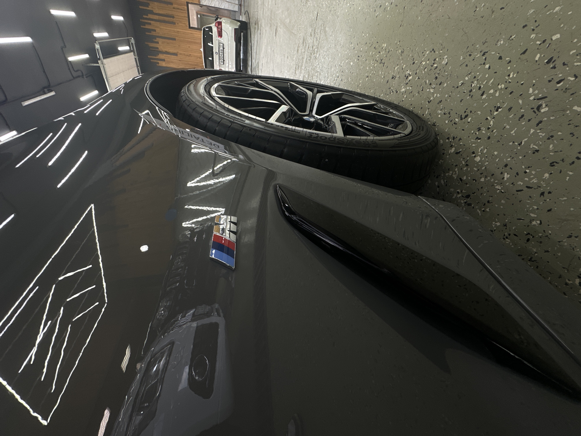 Here is a forensic description of the image.
**Scene:** The image captures a close-up view of a gray car in a detail shop or garage.
**Objects:** The primary objects are the car's wheel, tire, side panel, and an "M" badge. The wheel features a complex spoke design. The tire is black. The "M" badge is rectangular and shows red, blue, and navy colors from top to bottom.
There is a reflection of a white car and a stand on the car’s surface. The floor is speckled with an off-white color.
**Environment:** The location appears to be an indoor detailing shop or garage, indicated by the bright artificial lighting reflecting off the car's surface. The floor has a speckled epoxy finish.
**People/Subjects:** There are no people in the view of the image.
**Actions/Interactions:** None are visible in the frame.
**Overall Impression:** The scene suggests a car detailing or maintenance setting, with a focus on the car's exterior elements.
