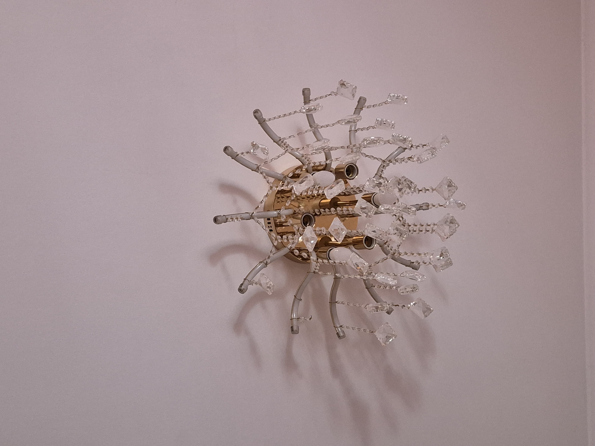 The image displays a decorative wall light fixture (sconce) mounted on a plain, light pink or lavender-colored wall. The fixture features a central, circular golden-toned base from which multiple curved, light grey or white arms radiate outwards. Attached to these arms and surrounding the central base are numerous clear, faceted crystalline or glass elements of various shapes and sizes, some strung together with smaller beads, creating a starburst or spiky appearance. Several small, tubular light bulb sockets are visible within the golden base.
Distinct shadows are cast on the wall behind and around the fixture, suggesting illumination from the fixture itself or an external light source. The wall surface is smooth with a subtle texture. No people or specific activities are depicted. The scene is an interior setting, likely a room or hallway. The ornate design of the light fixture evokes a vintage or classic aesthetic. No visible text is present in the image. The location context for this interior scene is Tobolsk, Russia.