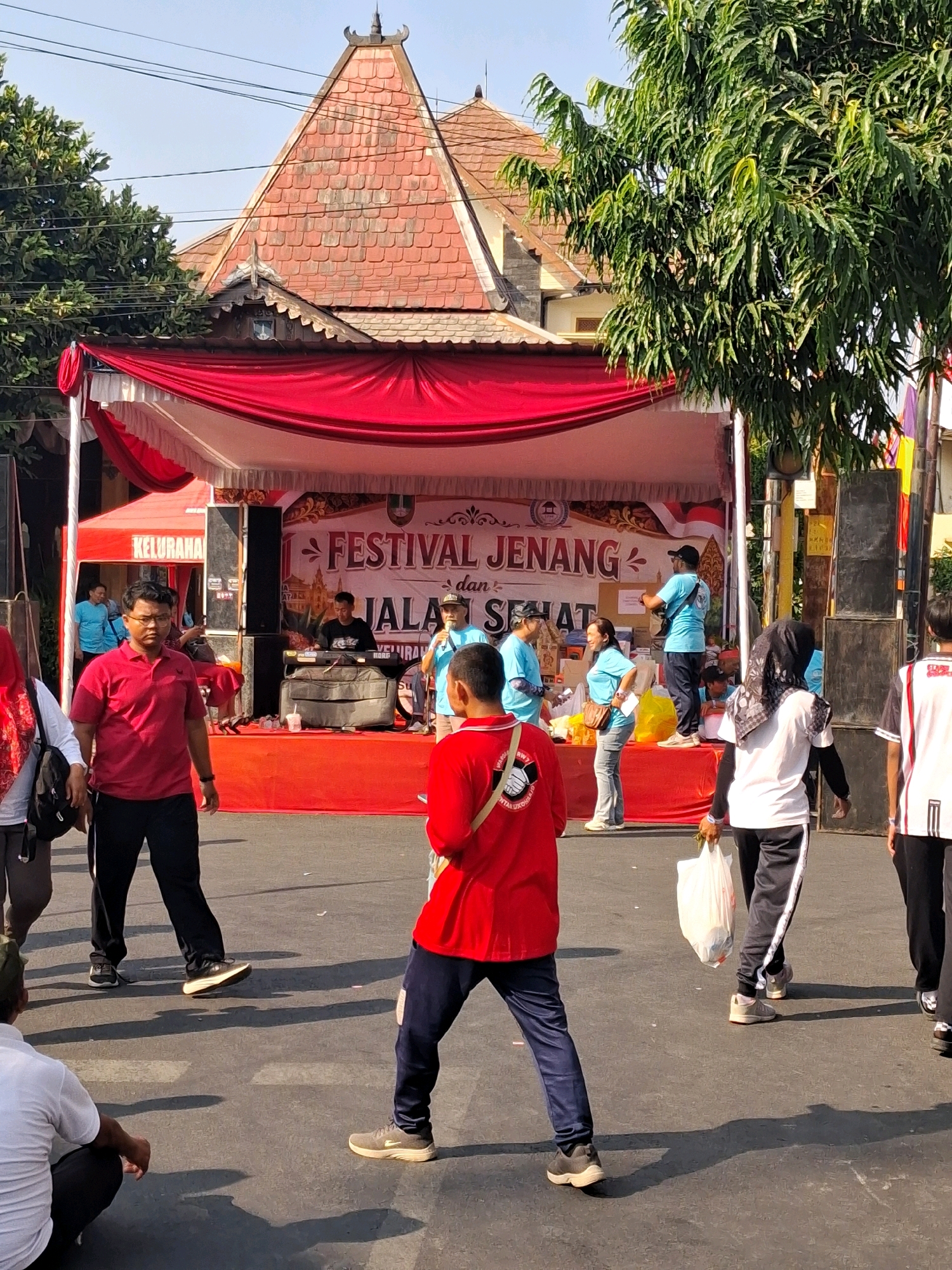 The image depicts an outdoor festival, likely in Surakarta, Indonesia, given the architectural style of the building in the background and the banner text which appears to be in Indonesian. The scene is set on a street with a stage erected for a performance or event.
In the foreground, a person in a red polo shirt and black pants is walking towards the viewer, looking slightly left. To the left of this person, partially visible, is another individual in a red shirt and black pants, facing left. In the middle ground, several people are gathered around the stage. A man in a black t-shirt is operating a keyboard on the stage, while another man in a blue shirt is speaking into a microphone. Several other individuals in blue t-shirts are standing around the stage, some appearing to be event staff. A woman in a blue shirt and jeans is visible near a table with items on it.
The stage is draped with red fabric and a white canopy. Behind the stage, a building with a distinctive multi-tiered, red-tiled roof and ornate trim is visible, suggesting traditional Indonesian architecture. A large tree with dense green foliage stands to the right of the building. The ground is asphalt, and shadows of people and objects are cast across it, indicating a sunny day.
Further to the right, a person in a white t-shirt with black and white striped sleeves is walking away from the viewer, carrying a white plastic bag. Another person in a black and white shirt and hijab is also walking away. People are dressed in casual attire, and some appear to be wearing uniforms or event-specific clothing, particularly the individuals in blue. The overall atmosphere suggests a lively community event.