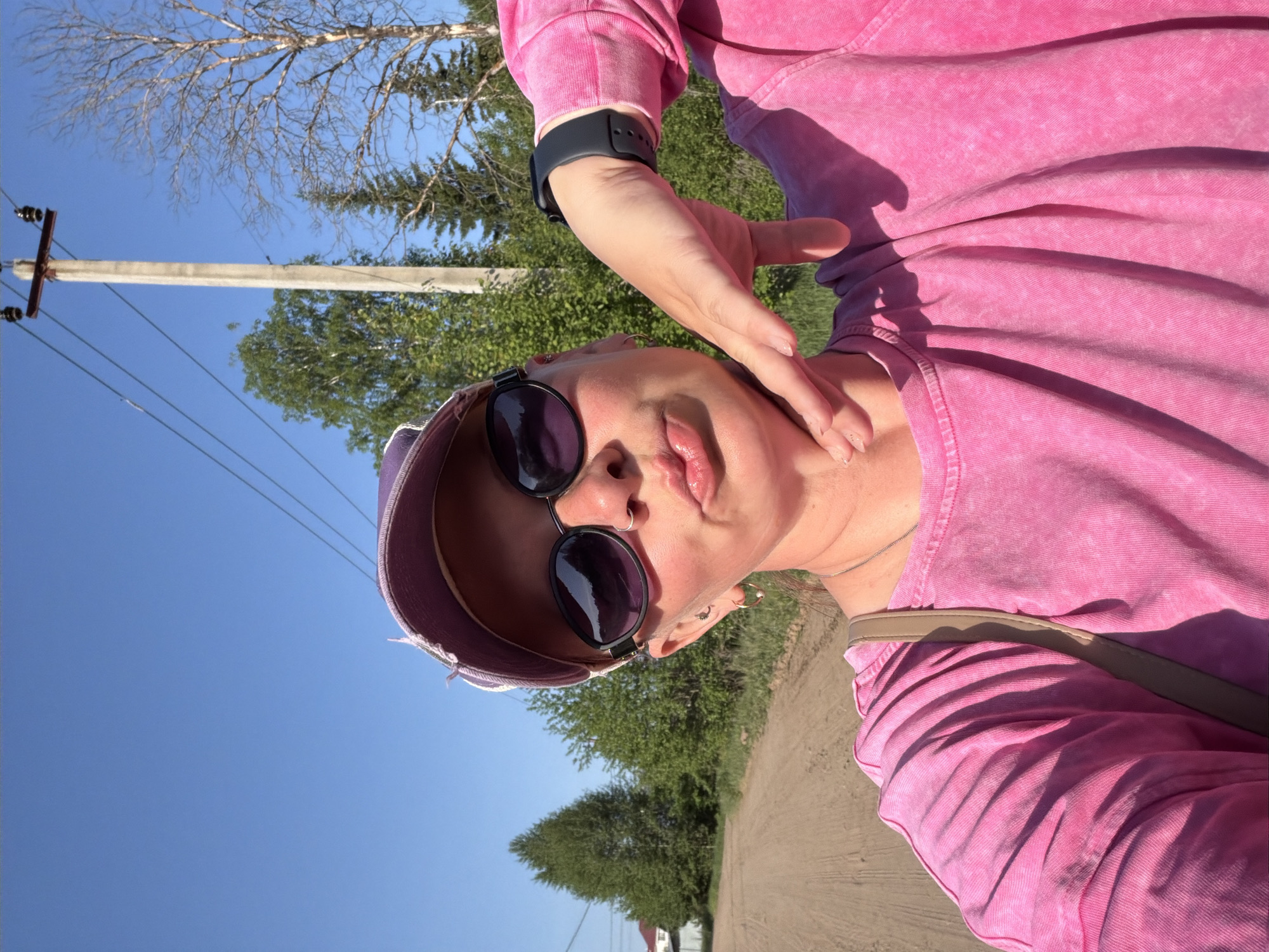 A person is taking a selfie outdoors on a sunny day in what appears to be a rural area. They are wearing a pink outfit, sunglasses, and a visor. The person is standing on a dirt path. In the background are green trees, a blue sky, and a utility pole. The location is the city of Chaikovsky, Russia.
