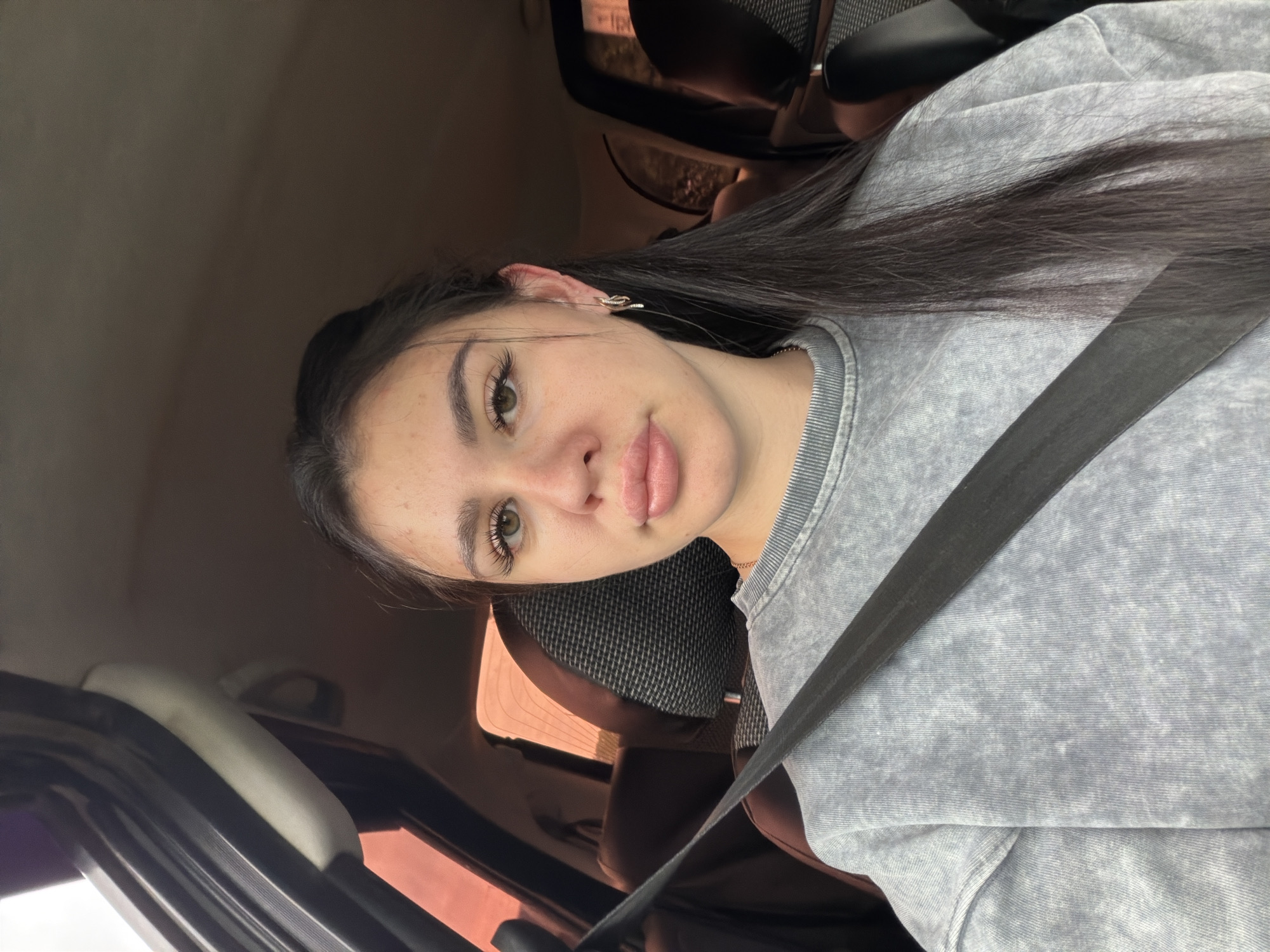 The image shows a young woman sitting in the driver's seat of a car, taking a selfie. She has long, dark hair and is wearing a grey sweatshirt and a seatbelt. Her makeup is natural, with emphasis on her lips and eyelashes. She has a lip ring and a snake-shaped earring in her left ear. The car's interior is visible, including the seats and windows, with some light suggesting it's daytime. The overall mood is casual and relaxed. The location is specified as Фоки, Russia.
