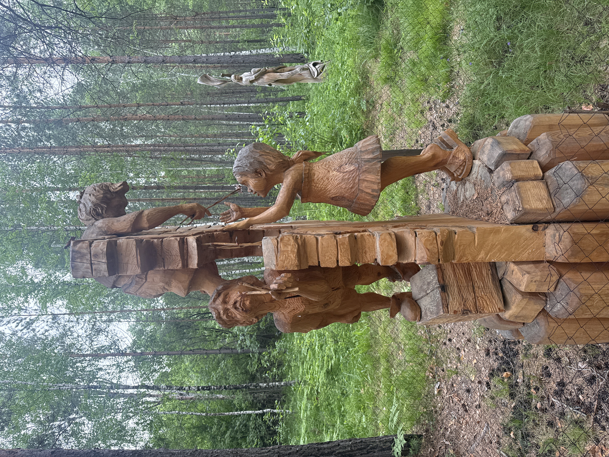 This image depicts an outdoor wooden sculpture located in the city of Savvateevka, Russia. The sculpture features three figures: an adult male and two children, a boy and a girl. The male figure, carved from wood, is positioned behind the boy, appearing to be supporting a large, segmented wooden structure above them. The boy, also carved from wood, stands to the left of the structure, looking upwards with an outstretched hand. The girl, a smaller wooden carving, stands to the right, reaching up towards the structure. The figures are intricately carved, with detailed facial features and clothing. The sculpture is set within a forest environment, with tall trees and lush green foliage surrounding it. A wire fence is visible in the foreground, partially obscuring the base of the sculpture. In the background, another wooden sculpture of a standing figure can be seen among the trees. The ground is covered with pine needles and grass.