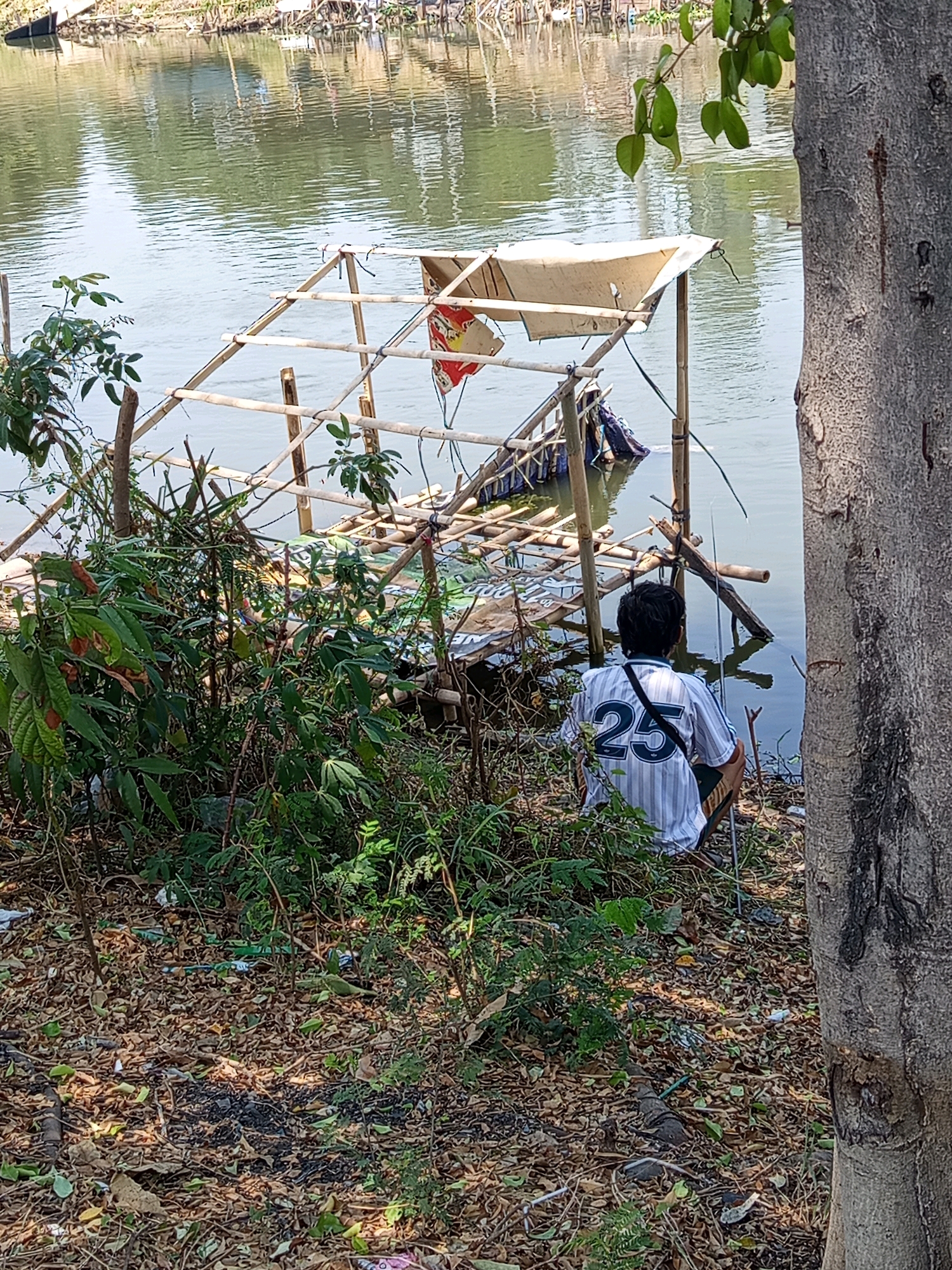 A person wearing a striped shirt with the number 25 is squatting near a body of water, possibly a river, in Karawang, Indonesia. The person is facing away from the camera and appears to be fishing, with a fishing rod visible. The area around the person is overgrown with vegetation and littered with debris, including leaves, plastic, and what appears to be remnants of a makeshift shelter constructed from bamboo. In the background, the water reflects the surrounding environment, and a dense collection of bamboo structures is partially submerged in the water. The overall scene suggests an informal settlement or a neglected area along the waterway.