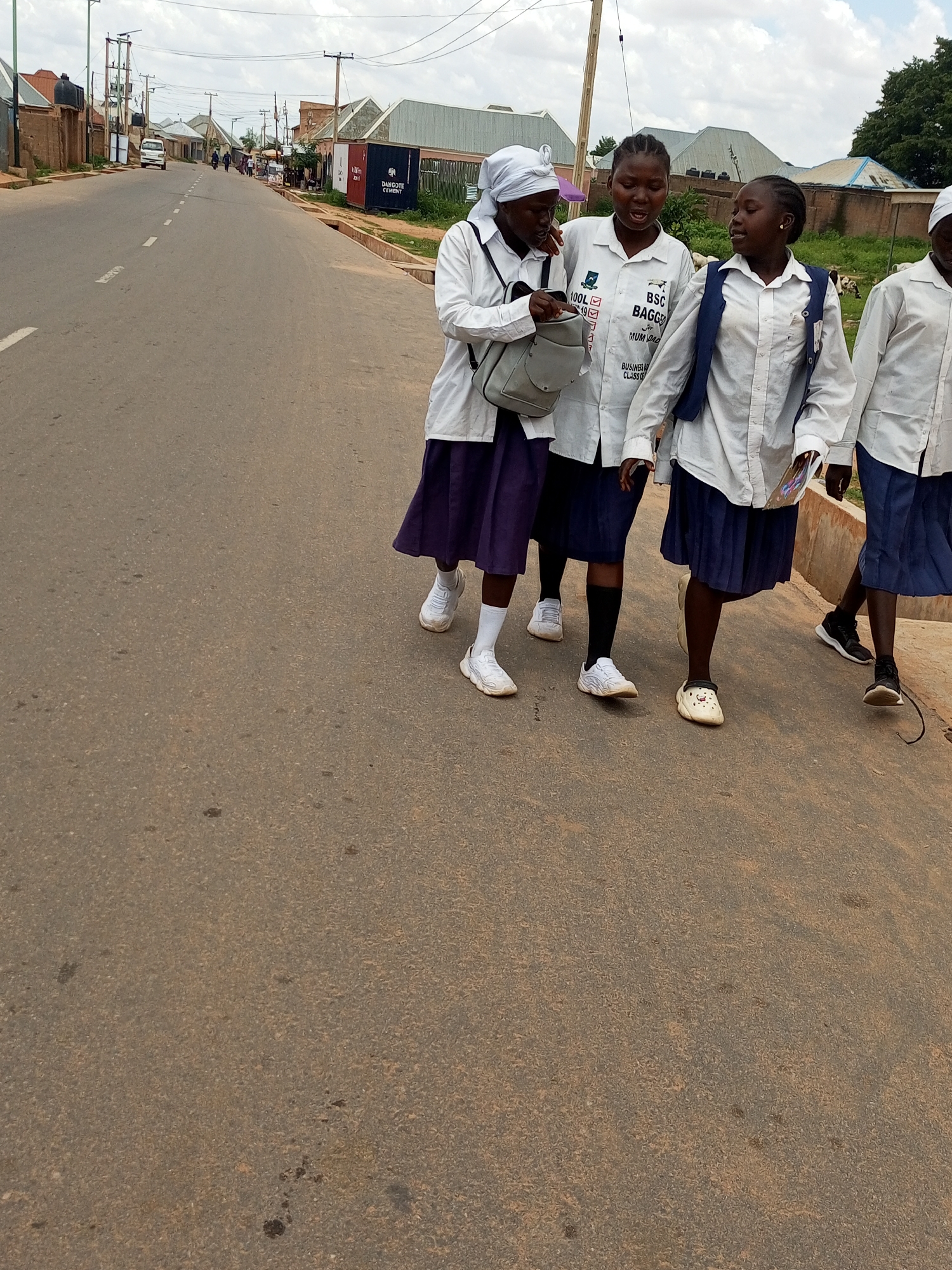 The image shows four young females walking on a paved road in what appears to be a Nigerian town, identified as Tempure. They are dressed in school uniforms consisting of white shirts and dark-colored skirts, with some wearing headscarves. The girls are walking in a group, engaged in conversation. In the background, there are buildings, utility poles, and some vegetation. The road is a typical urban street with visible lane markings. The sky is overcast, suggesting a cloudy day. The overall scene depicts a normal day in a Nigerian community, with the students walking together.