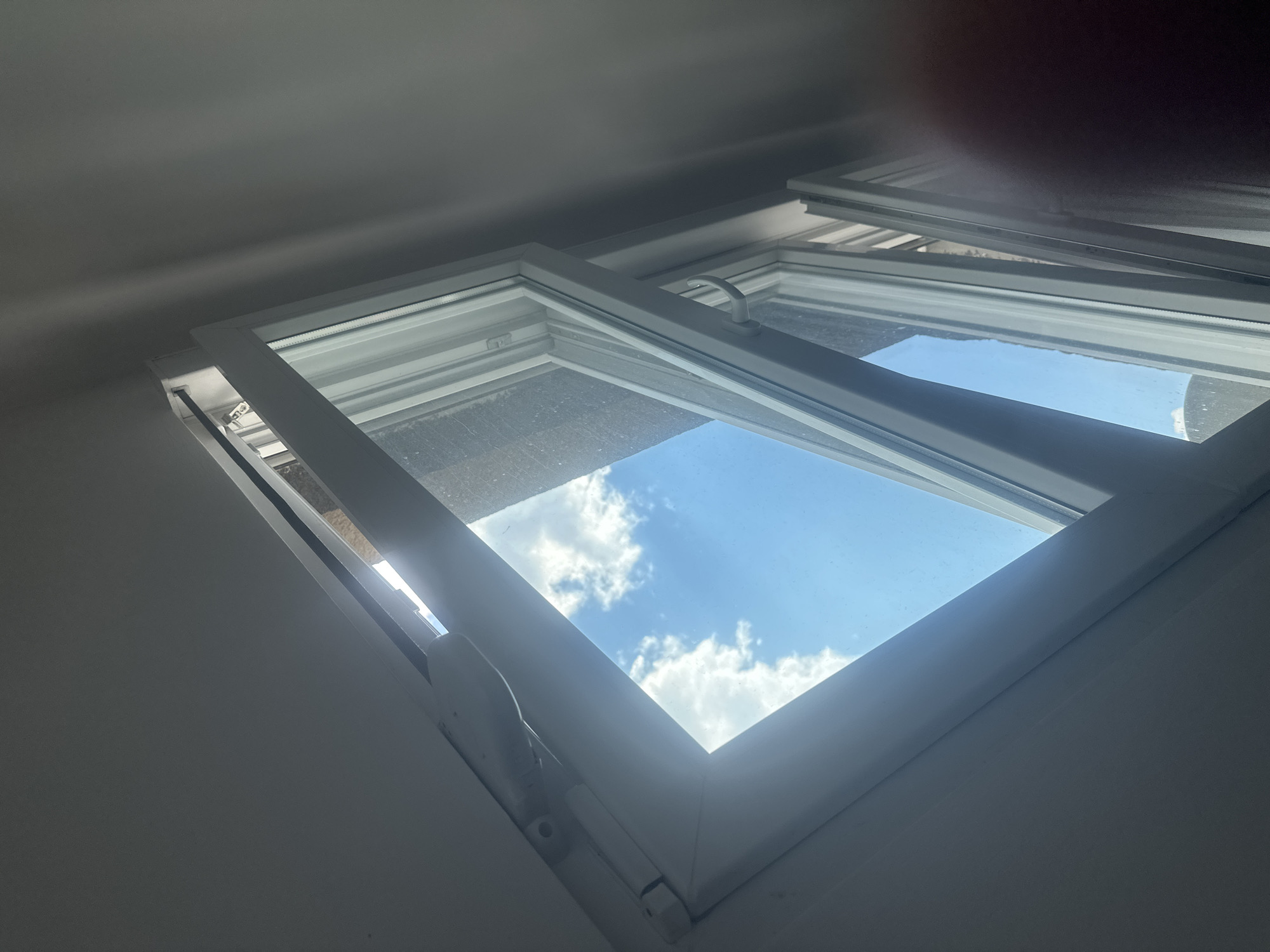 The image shows an interior view of a white-framed skylight, tilted open to reveal a bright blue sky with fluffy white clouds. Sunlight streams through the opening, casting shadows on the white interior walls. The window appears to be a casement window, with hinges visible along the bottom edge. A white handle is used to operate the window. The overall scene suggests a clear day, likely daytime, given the brightness of the sky and the sun's illumination. The setting is an indoor space with white, angled walls, possibly a room with a pitched roof or attic. There is no visible text in the image. The overall mood is one of openness and natural light.