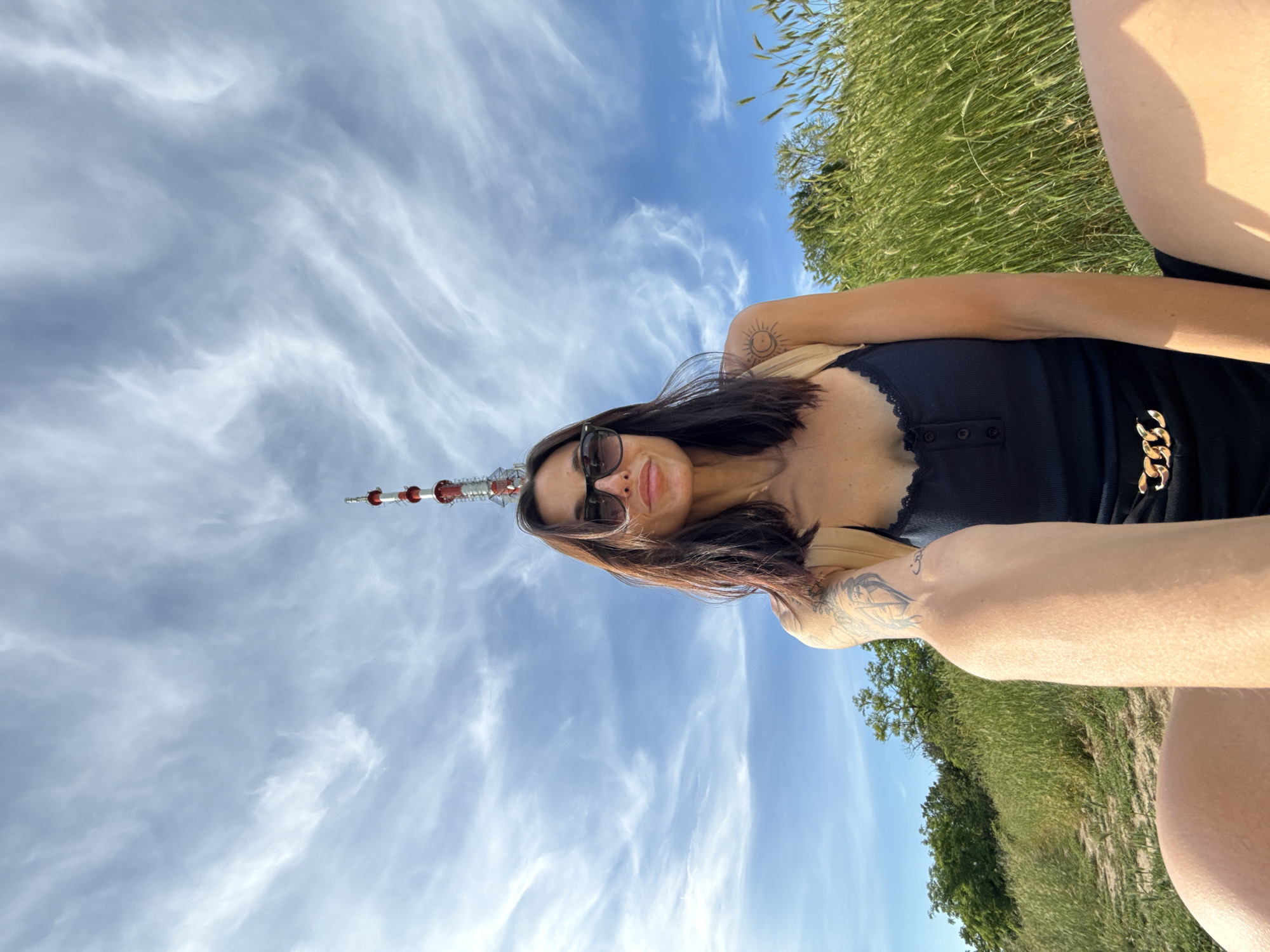 A woman is taking a selfie outdoors on a sunny day, with a red and white telecommunications tower visible in the background, centered above her head. She has dark hair, sunglasses, and is wearing a black tank top and black shorts with a gold chain detail. She has tattoos on her arm and shoulder. The environment appears to be a grassy hilltop with trees in the distance. The telecommunications tower is located on the Pleven Ridge, a historically significant area in Pleven, Bulgaria.
