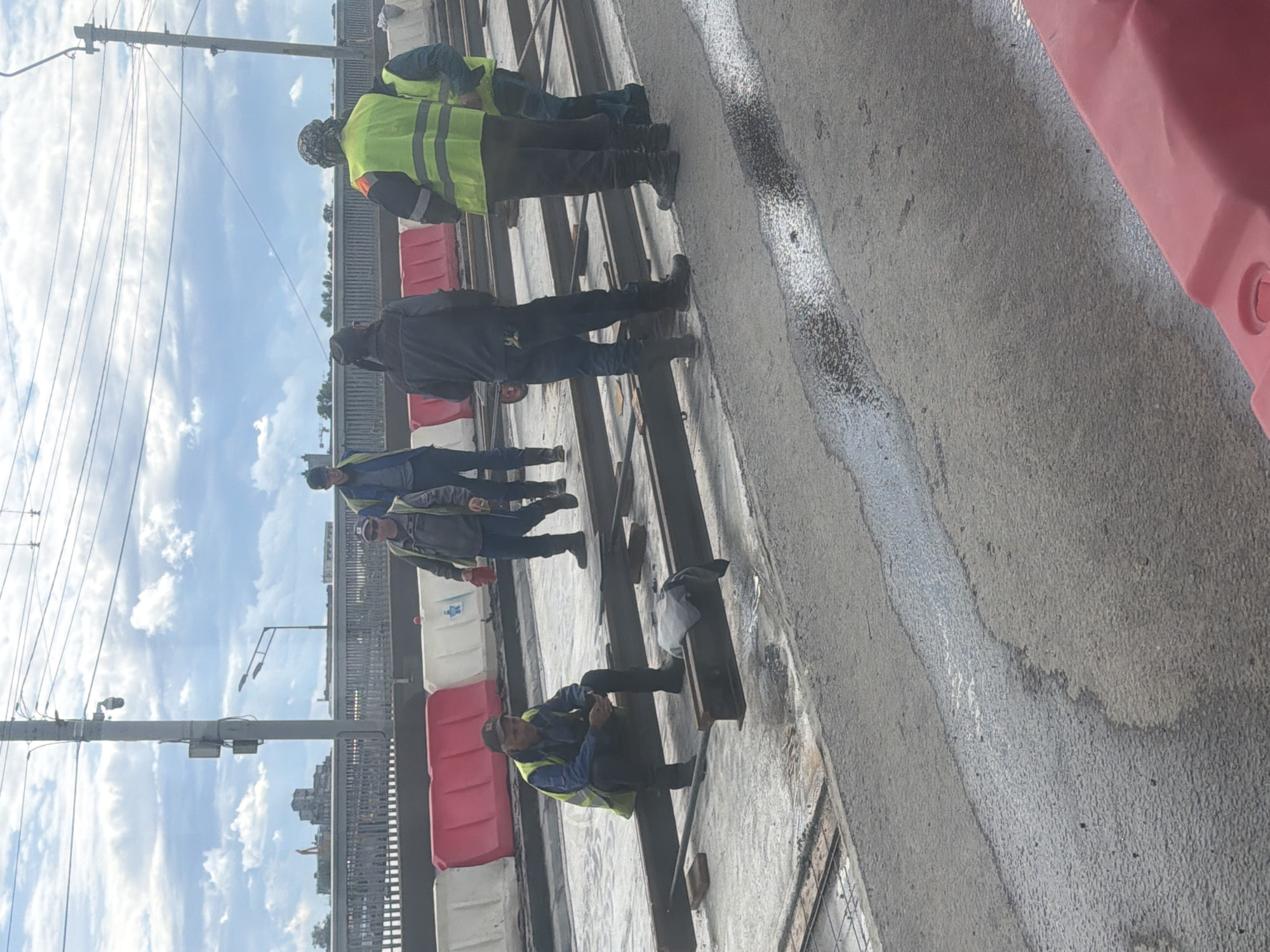The image shows a group of construction workers on an outdoor site, likely in Saint Petersburg, Russia. The sky is partly cloudy, suggesting daytime. The workers are wearing high-visibility vests and workwear, indicating they are engaged in some form of construction or infrastructure maintenance. One worker is crouched down on a metal beam, appearing to be taking a break or working on something. Others are standing and observing. The scene includes construction barriers, metal structures, a utility pole with wires, and a cloudy sky with some blue patches. The overall impression is of a typical workday on a construction project. There is no visible text in the image.