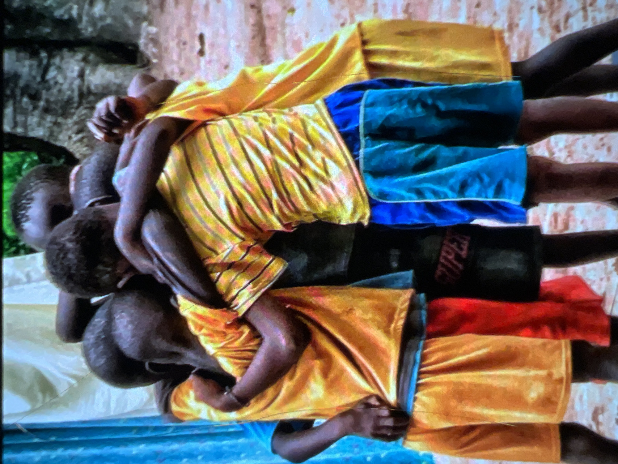 A group of four young Black boys are in a close embrace, their bodies intertwined in the center of the frame. They are all wearing brightly colored shorts and t-shirts. The boy on the far left wears a yellow t-shirt and orange shorts. Next to him, a boy in a yellow and orange striped t-shirt and blue shorts. The third boy, partially obscured, wears a black t-shirt with white lettering, possibly a logo. The boy on the far right wears a yellow t-shirt and yellow shorts. Their skin tones are dark. The background consists of a sandy ground, trees, and a light-colored cloth draped on the left side, possibly a curtain or a tent. The overall impression is one of warmth and camaraderie.