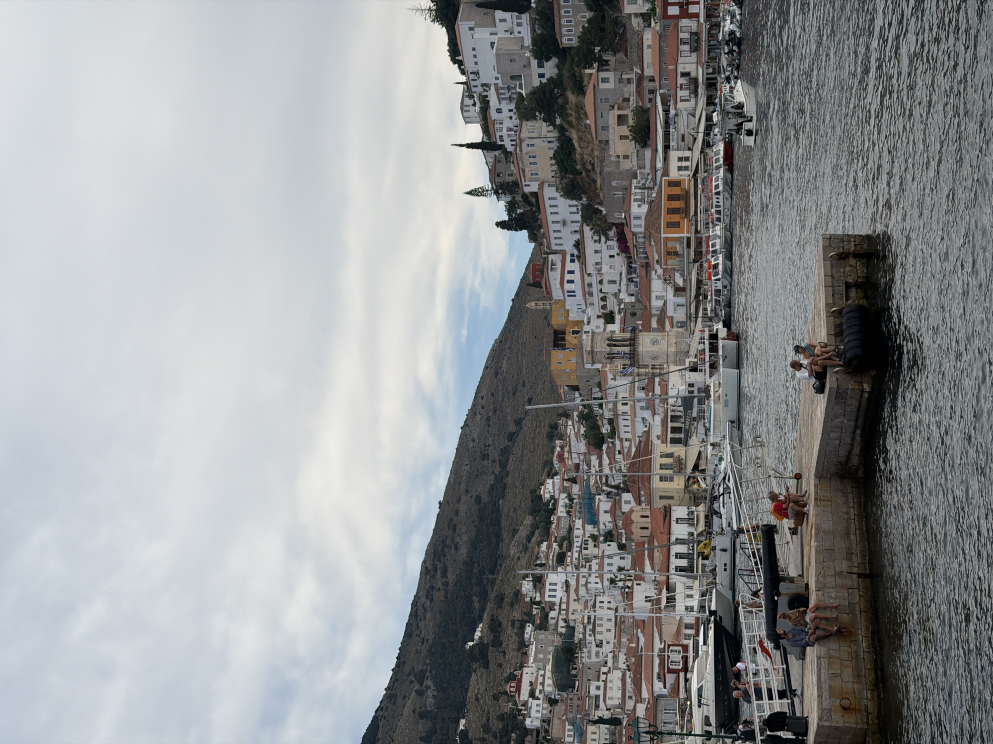 The image shows a view of the town of Hydra, Greece, as seen from the waterfront. The town is built on a hillside, with whitewashed buildings with red-tiled roofs. A mountain rises on the left side of the image. The sea is on the right. Several people are sitting along the waterfront and harbor-side. There are boats docked along the harbor. The sky is cloudy.