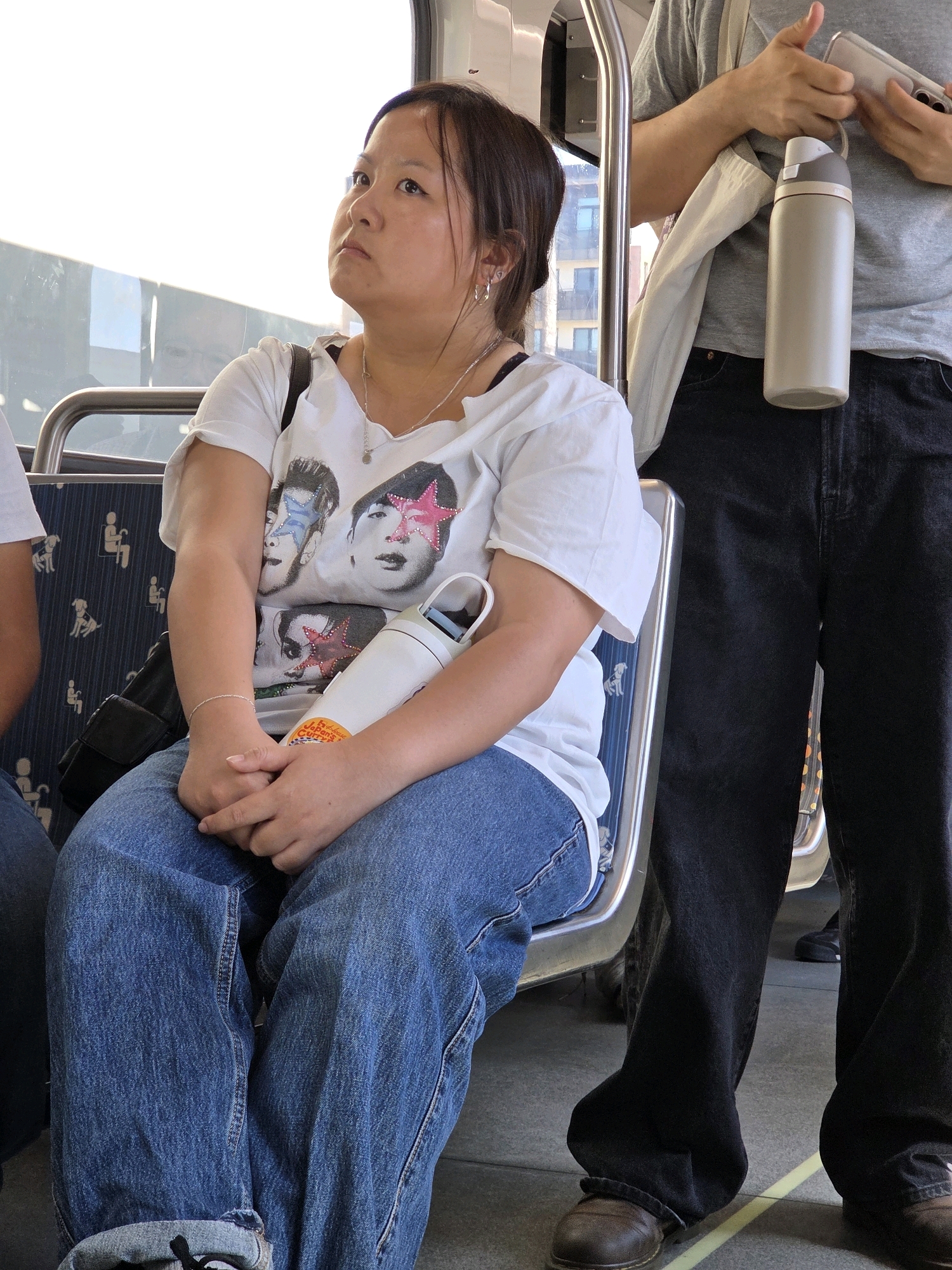 A woman sits on a public transport seat, her legs crossed, wearing blue jeans and a white t-shirt with an image of David Bowie on it. A water bottle with a white body and a grey lid sits on her lap, alongside her folded hands. The seat she occupies has a blue pattern with white figures depicting people and dogs. Beside her, a person wearing black jeans and a grey t-shirt holds a phone and a light-colored water bottle. The floor of the transport is grey, with yellow lines indicating spacing. The scene is set in an urban environment, likely on a bus or train, given the seating arrangement and handrails.