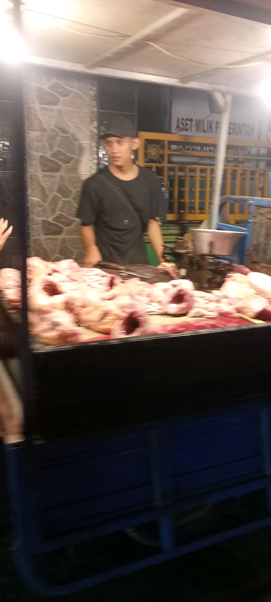 The image shows a street vendor at a market stall in Singogalih, Indonesia. A young man wearing a black t-shirt and a black baseball cap stands behind a display of raw chicken. A large cleaver rests on the counter in front of him. The stall is illuminated by overhead lights. In the background, there is a sign that reads "ASET MILIK PEMERINTAH" which translates to "Government Property". The stall appears to be outdoors, with a stone wall and a metal fence visible behind the vendor. The ground is covered in dark material.