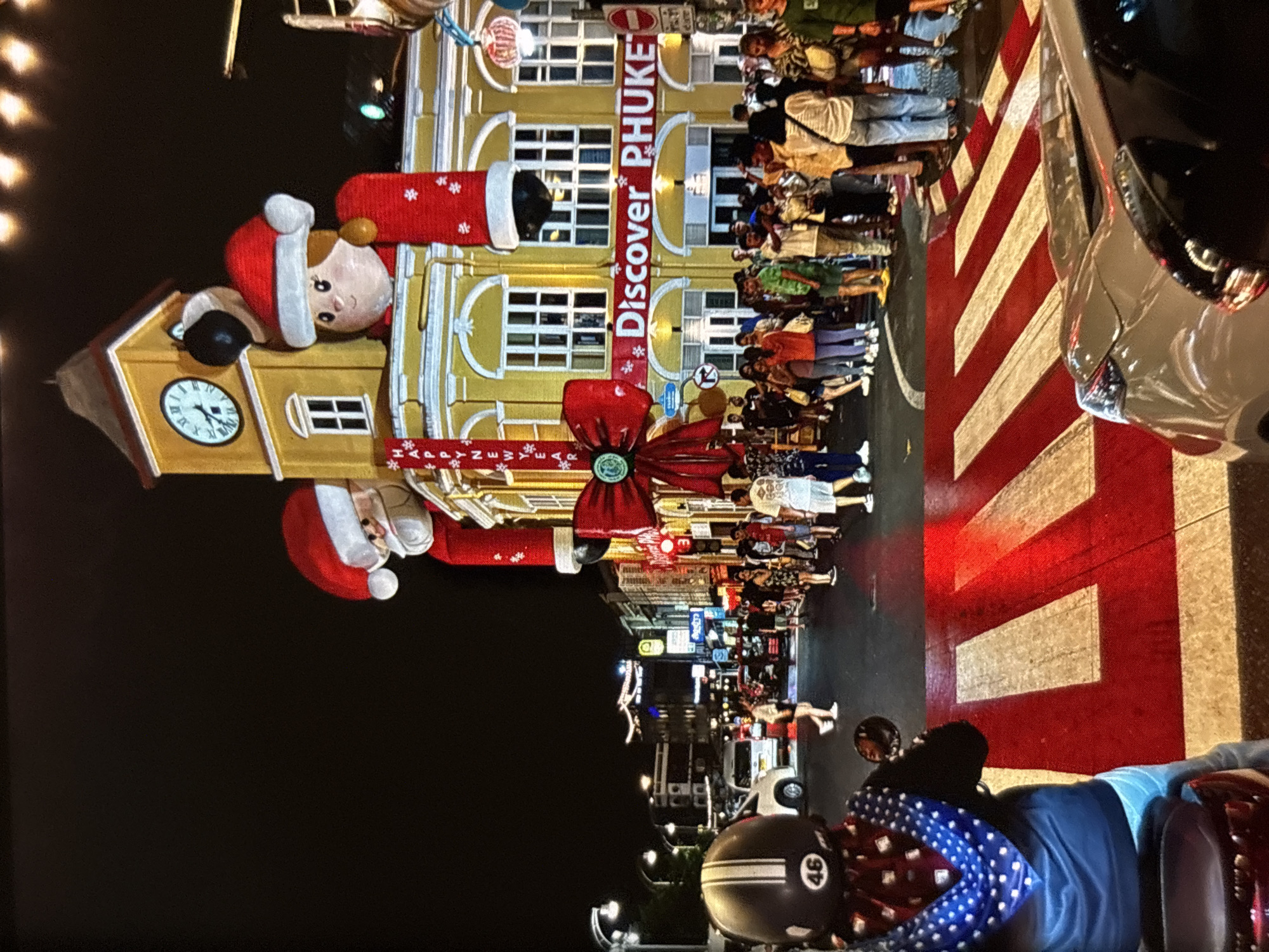 The image captures a festive street scene in Chalong, Thailand, at night, likely during the Christmas and New Year season, as indicated by large inflatable Santa figures and "Discover Phuket" signage adorned with holiday decorations like a giant red bow and snowflake motifs. The focal point is a yellow building with a clock tower, decorated with these festive elements. A prominent crosswalk with red and white stripes is in the foreground, partially occupied by a silver car. On the left, a person on a scooter, wearing a helmet with "46" visible and a blue bandana, is positioned at the crosswalk. The street is bustling with a crowd of people, some of whom appear to be tourists, walking and gathered on the sidewalks. Streetlights and building lights illuminate the scene, creating a vibrant nighttime atmosphere. The background shows more buildings and streetlights, suggesting a commercial or entertainment district.