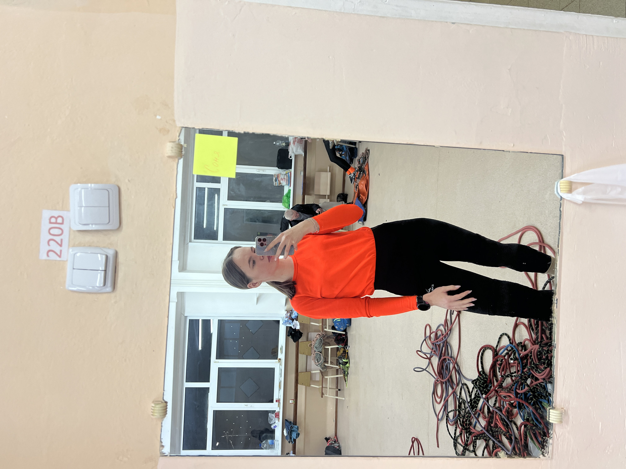 A young woman is taking a mirror selfie in a room. She is wearing a bright orange long-sleeved shirt and black leggings. The room appears to be a sports or training area. There are windows with multiple panes, wooden chairs stacked against the wall, and piles of climbing ropes on the floor. A handwritten note is affixed to the window frame in the upper left portion of the mirror's reflection. The room appears to be located in Berezniki, Russia. A light switch is visible on the wall next to the mirror, with the marking "220B."
