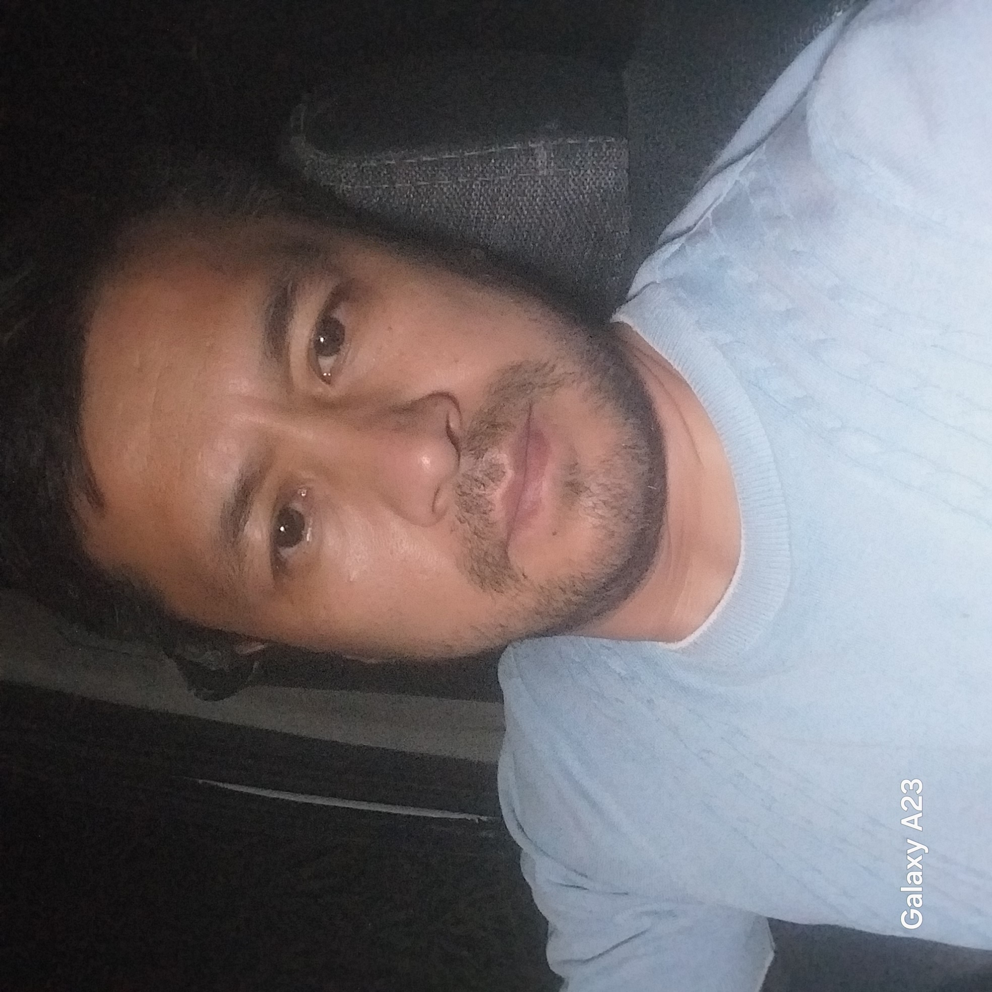 A close-up, waist-up, indoor, selfie-style photograph shows a man looking at the camera. The man is in his 30s or 40s, with short, dark hair, a medium complexion, and a light stubble. He is wearing a light blue crewneck shirt. The background is dark and out of focus, suggesting the photo was taken at night or in a dimly lit area. A watermark in the bottom left corner reads "Galaxy A23." The scene appears to be a vehicle interior, with the textured surface of a car seat visible behind the man.